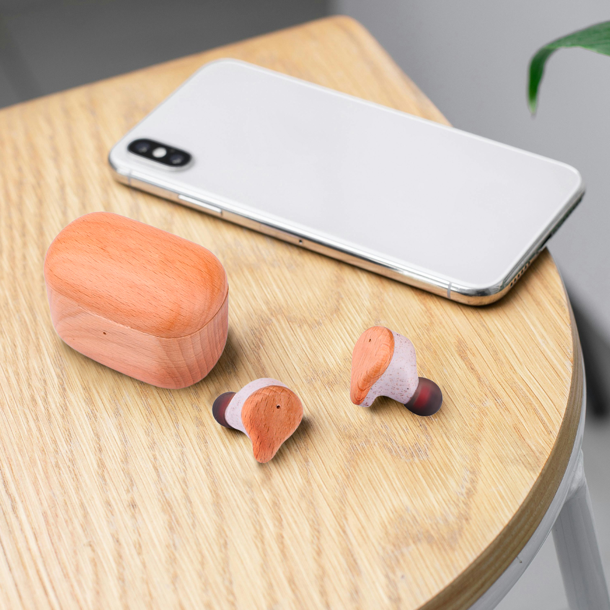 Auriculares de madera, estuche de carga y un teléfono blanco sobre una mesa de madera. Auriculares rosas y marrones.