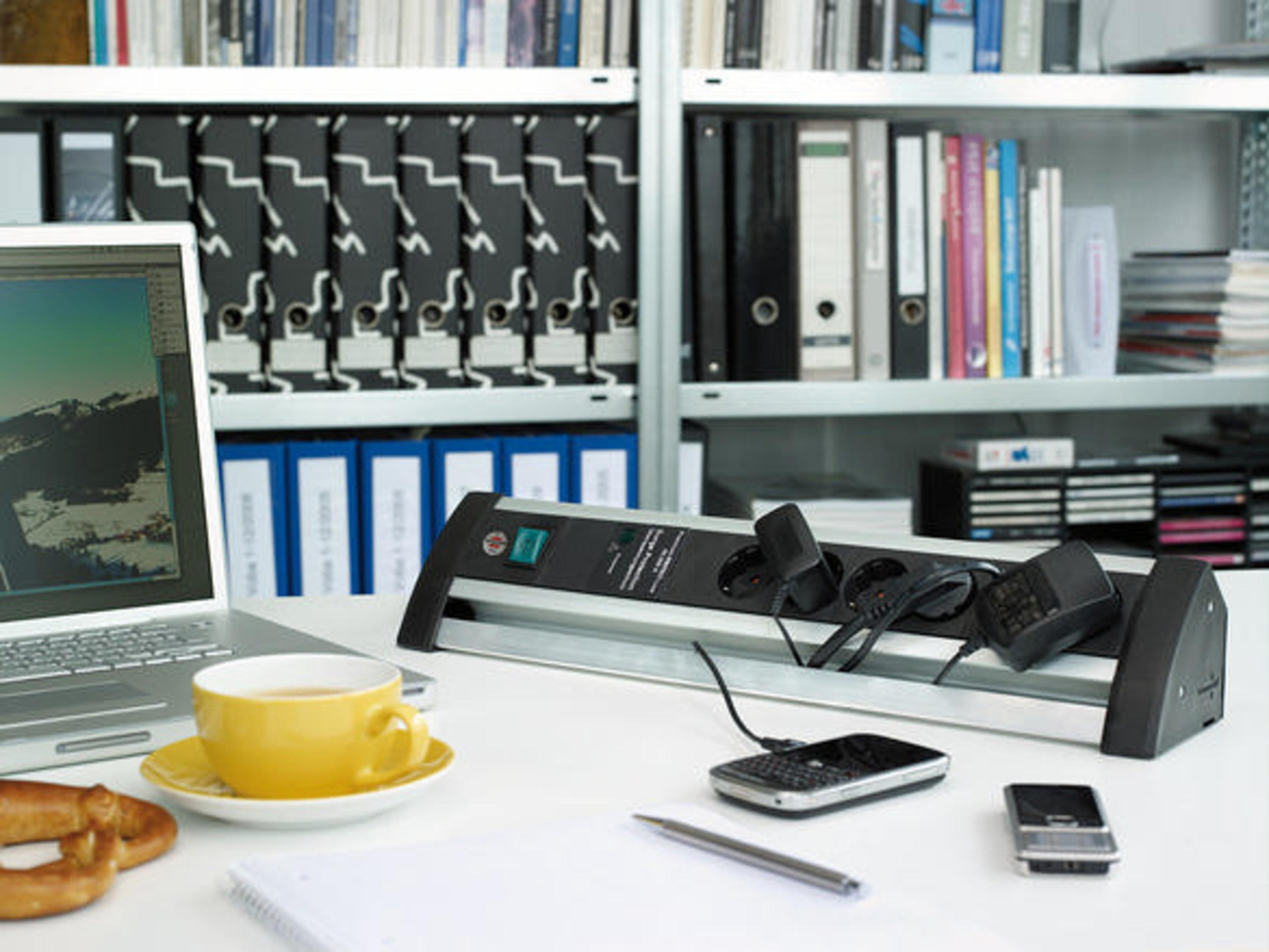 Dizüstü bilgisayar, telefon ve priz uzatma kablosu olan ofis masası. Sarı çay fincanı, kalem ve atıştırmalık. Arka plan kitaplık.
