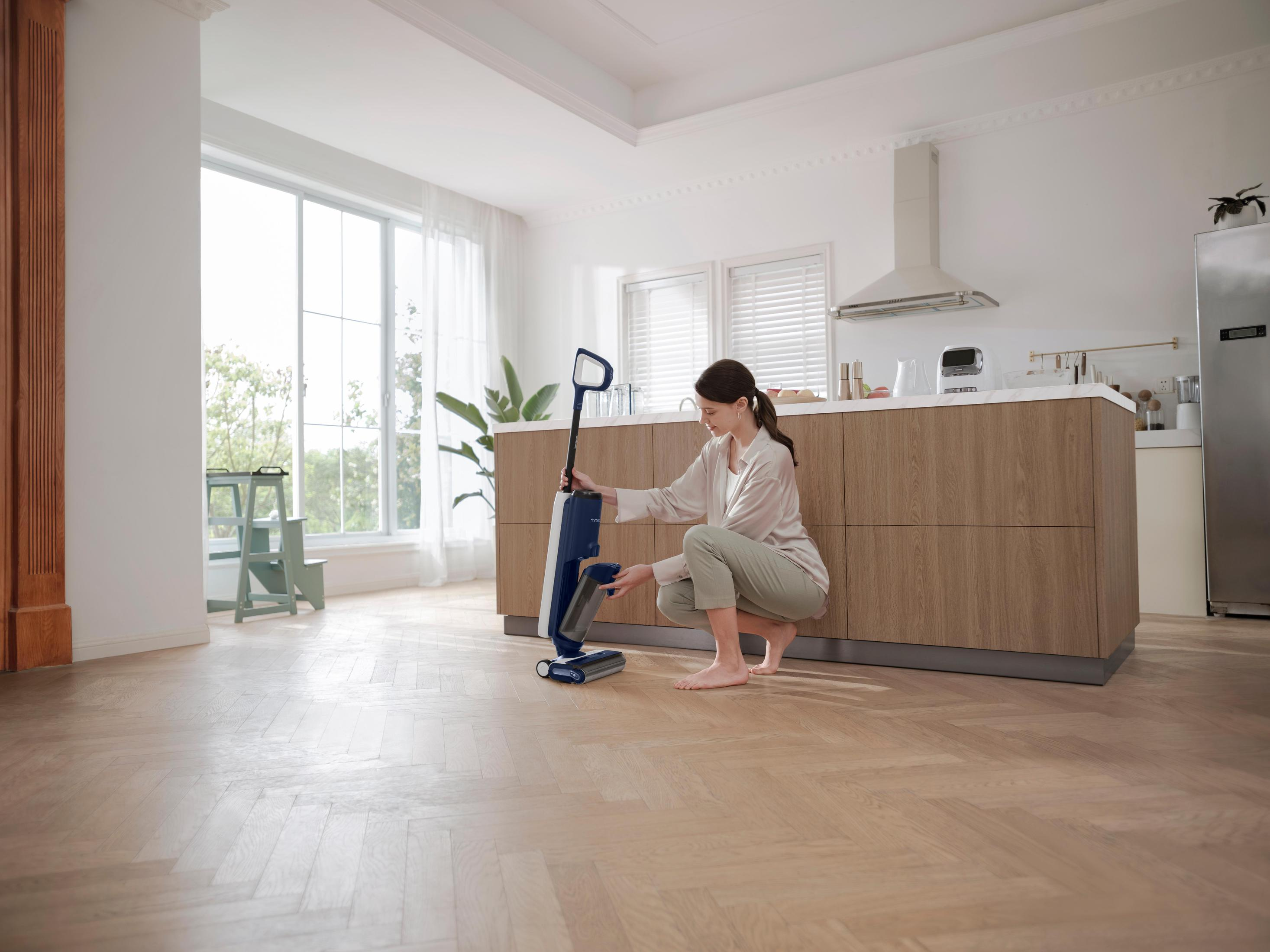 Kobieta używająca niebiesko-białego środka do czyszczenia podłóg w kuchni. Widoczna drewniana podłoga i szafki.