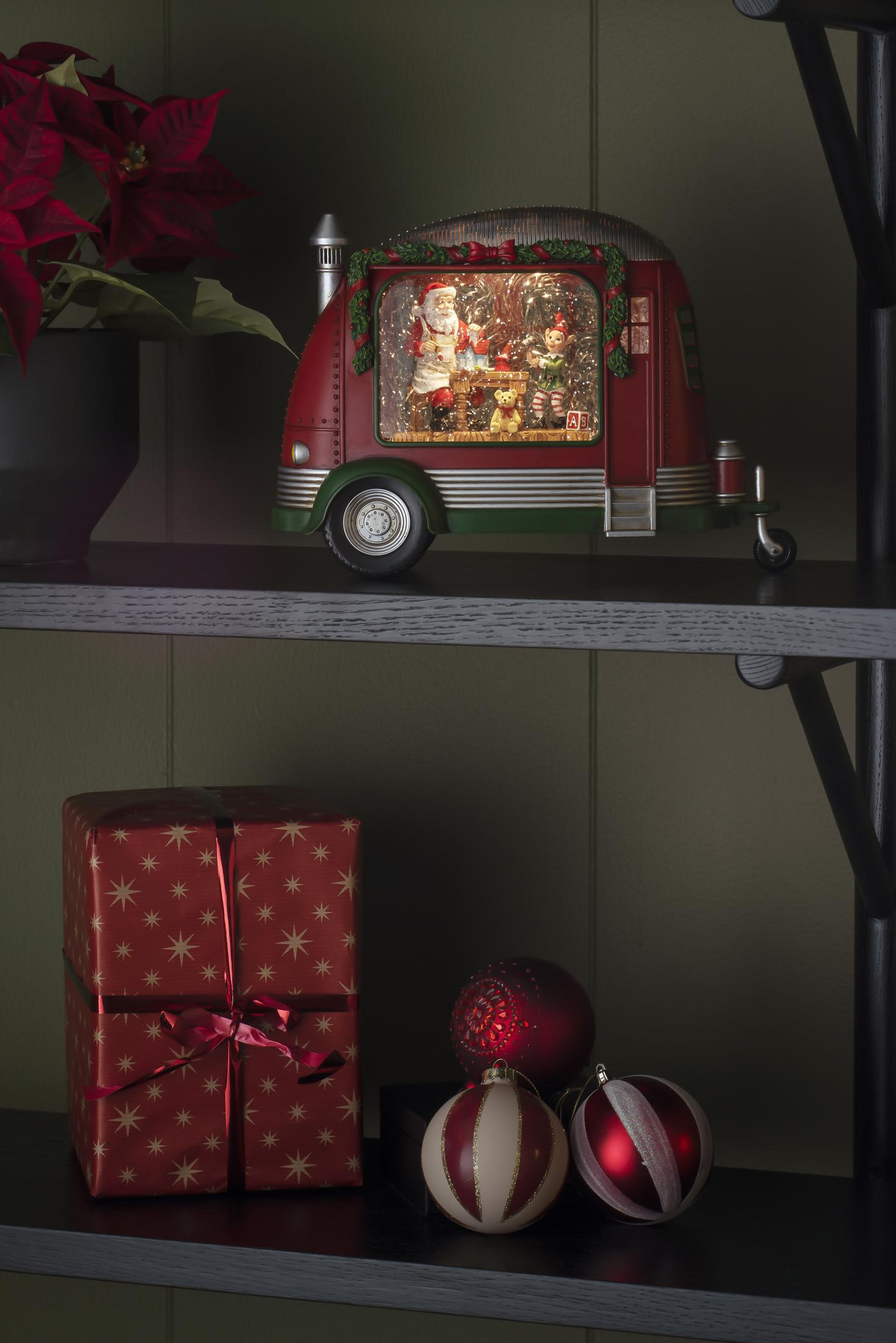 Weihnachtsszene mit rotem Wohnwagen, Weihnachtsmann darin und festlicher Dekoration auf einem Regal.
