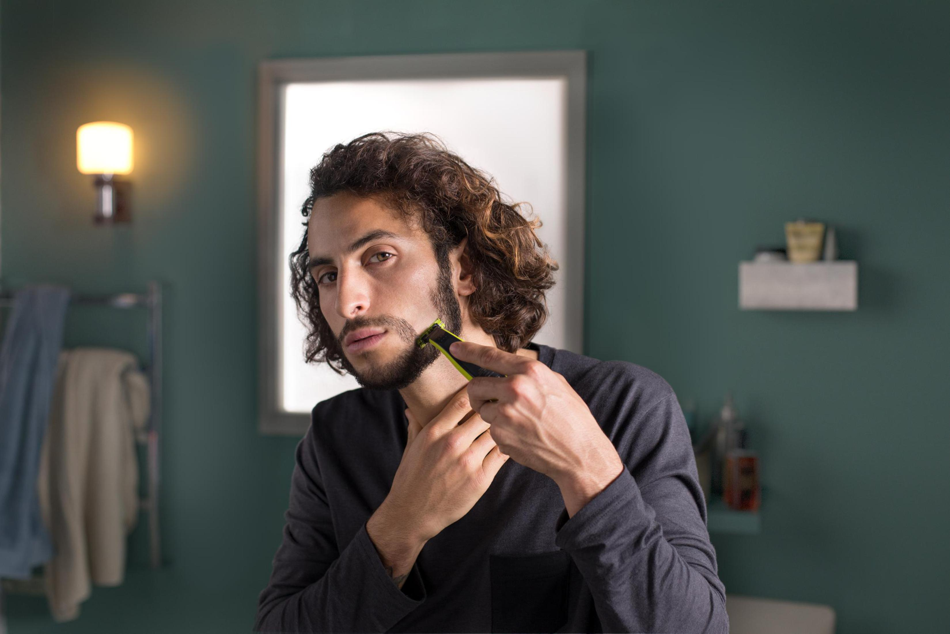 Mężczyzna goli brodę w łazience z lustrem, używając elektrycznej maszynki. Tło: ściana i światło.