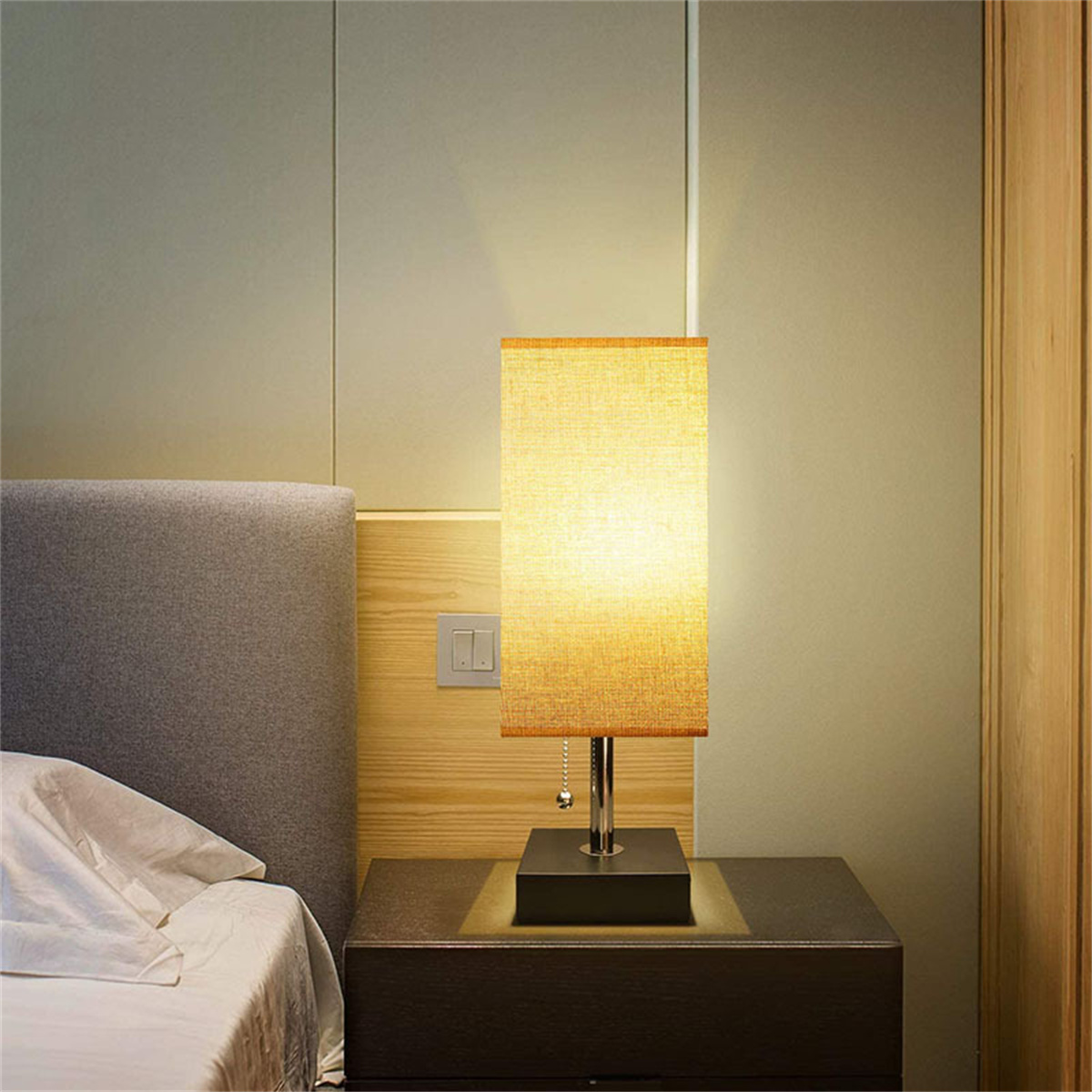 Eine Lampe steht auf einem Nachttisch. Ein Bett und eine Wand sind im Hintergrund.