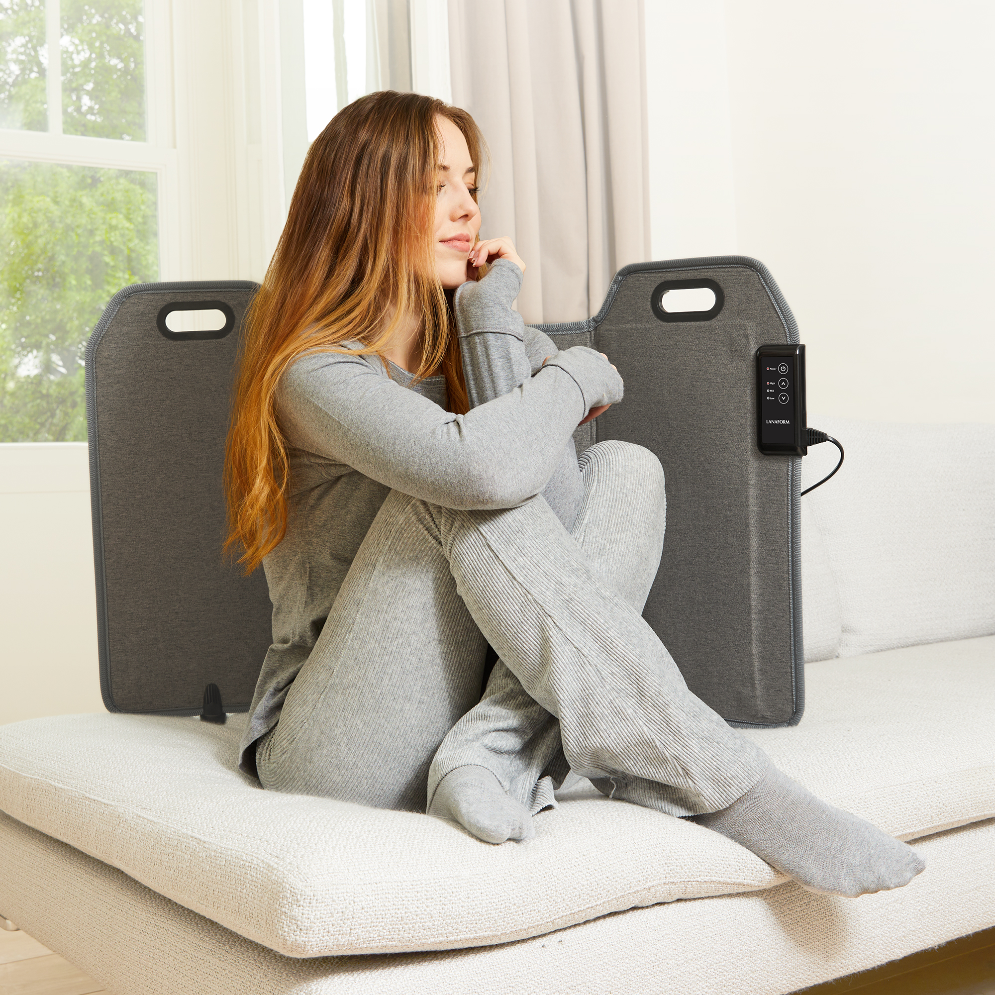 Eine Frau in grauen Jogginghosen sitzt auf einem Sofa mit einem grauen Heizkissen hinter sich, in der Nähe eines Fensters.