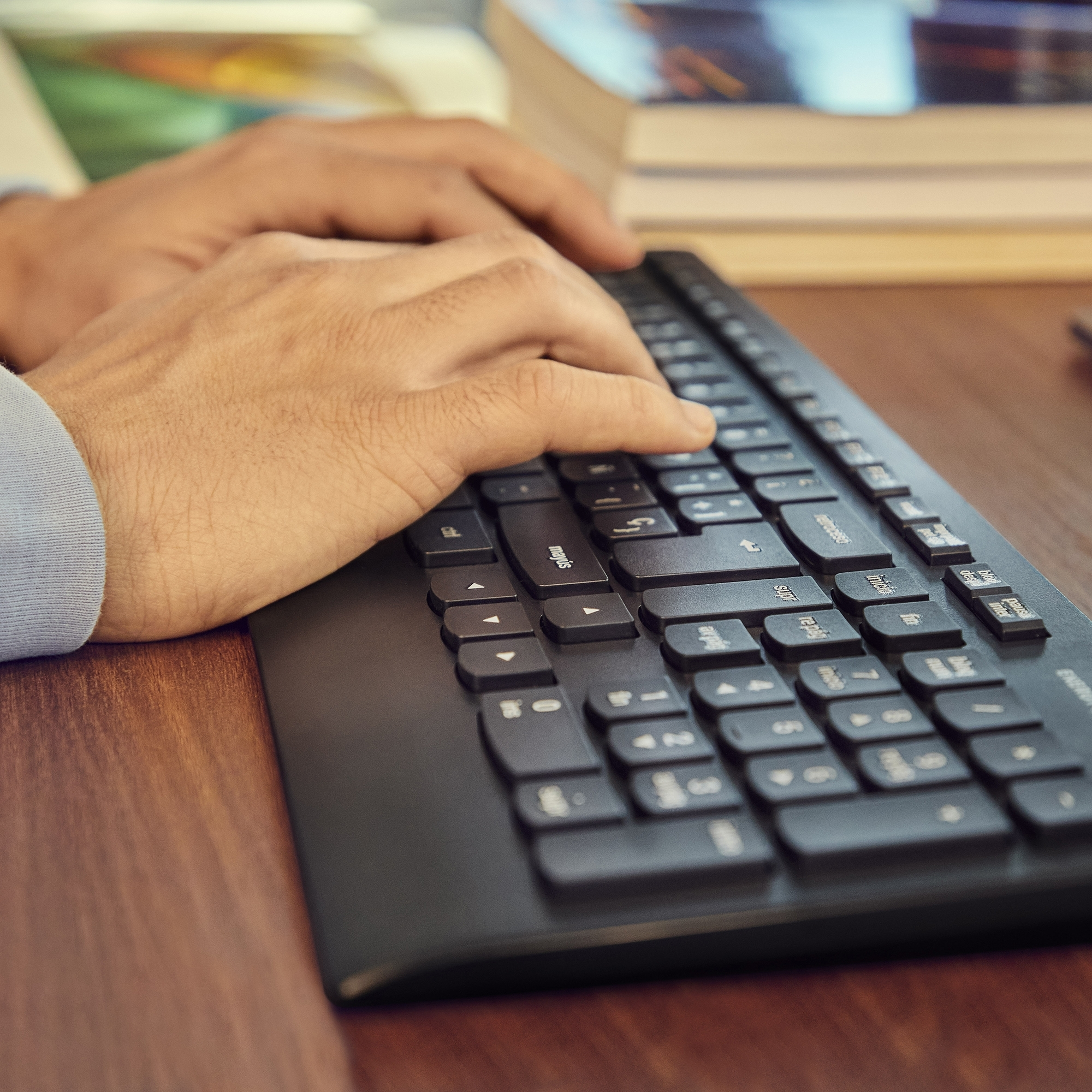 Manos tecleando en un teclado negro sobre un escritorio de madera, con libros al fondo.
