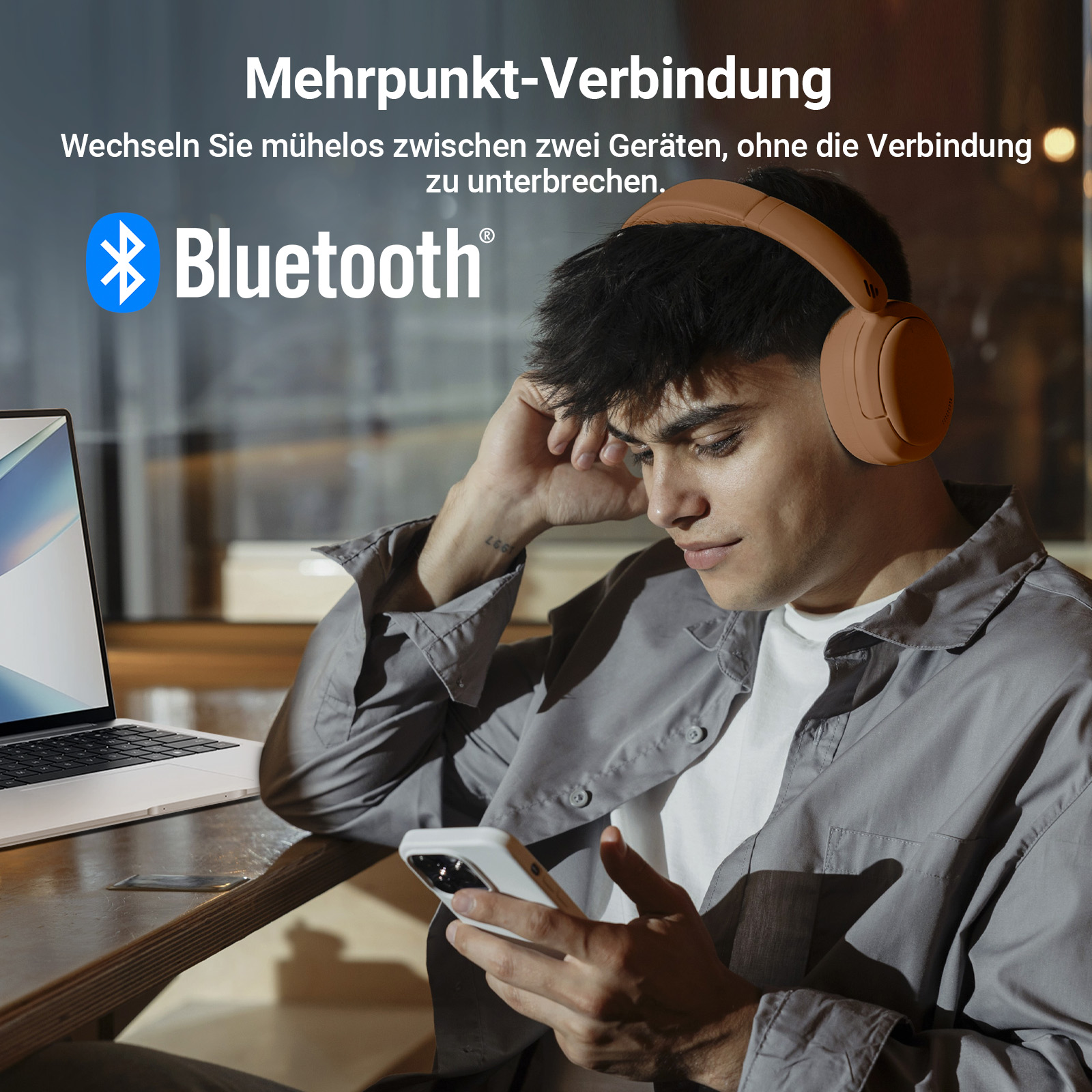 Ein Mann mit braunen Kopfhörern hält ein Telefon. Ein Laptop liegt auf dem Tisch.