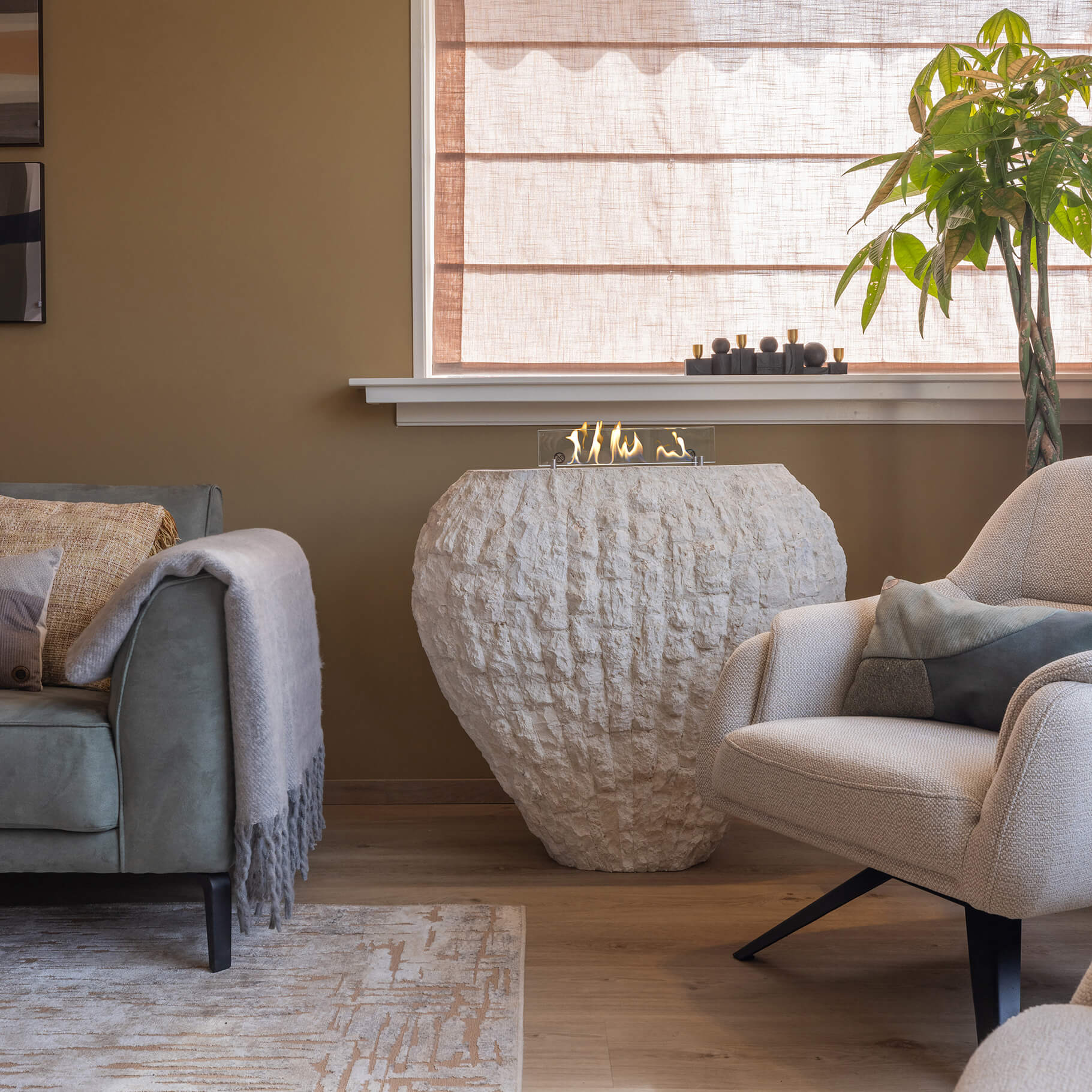 Een woonkamer met een bank, fauteuil en een decoratieve open haard. Een plant staat bij het raam.