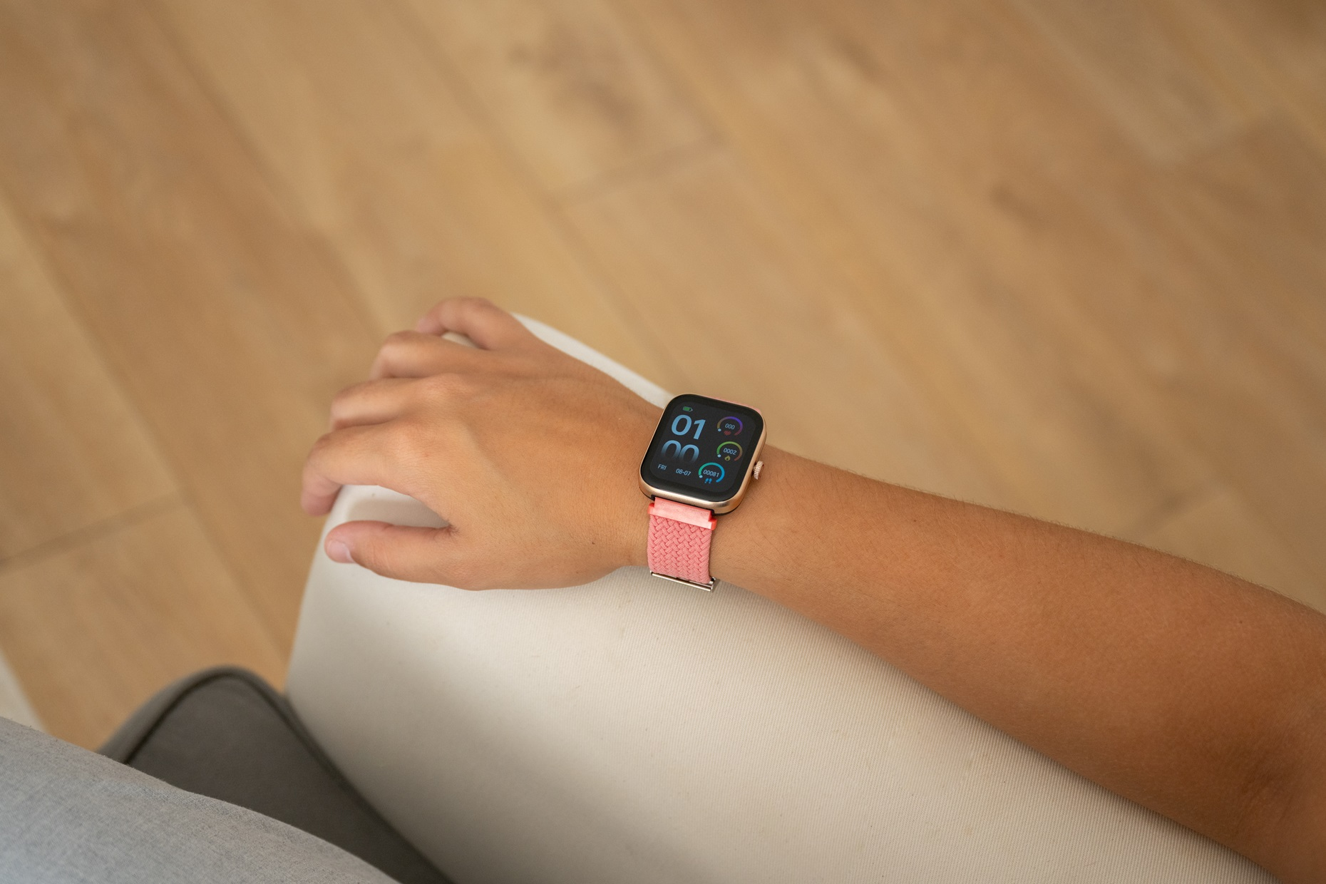 Brazo de persona con reloj inteligente rosa sobre fondo de madera clara, en interiores.
