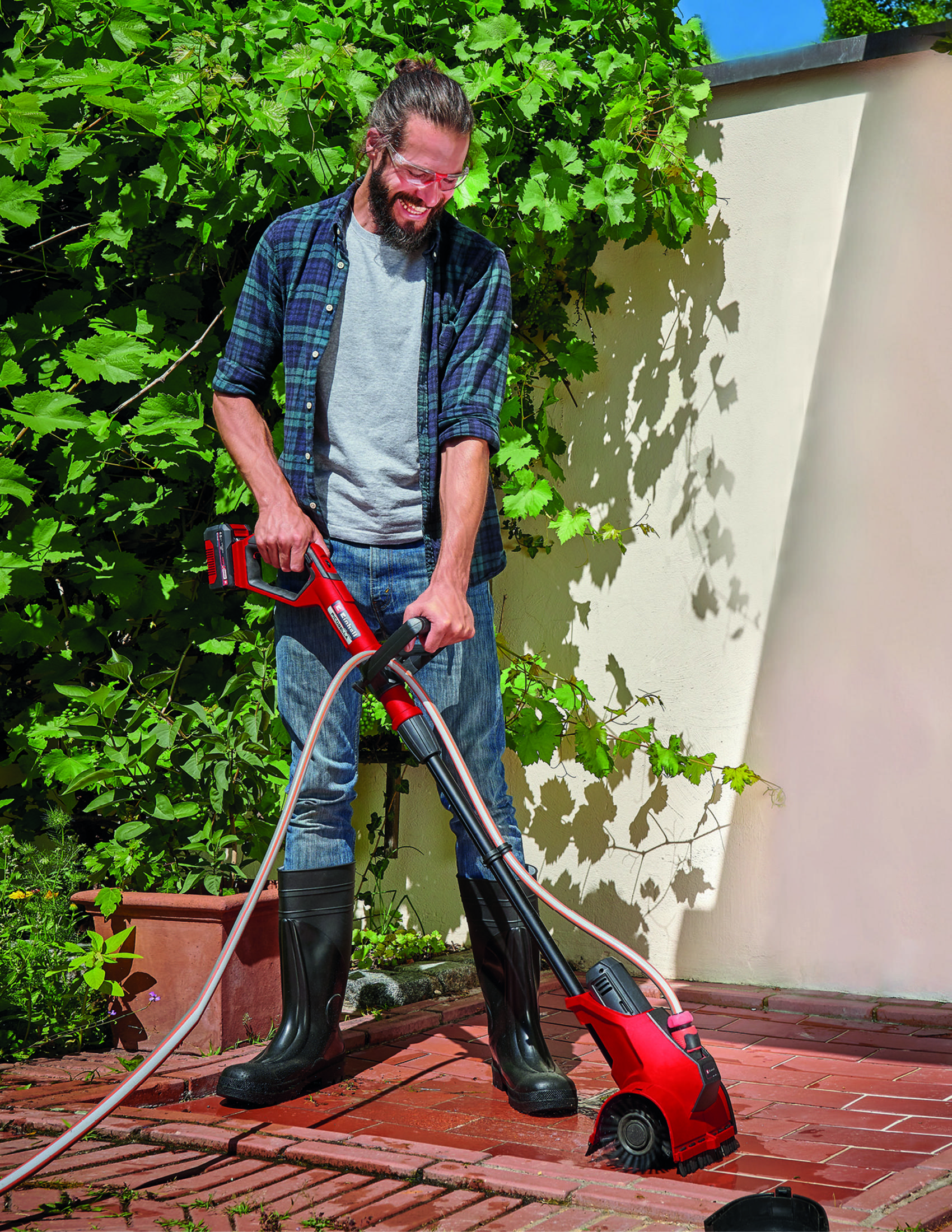 A man cleaning a brick patio with a red power washer, wearing boots and jeans.