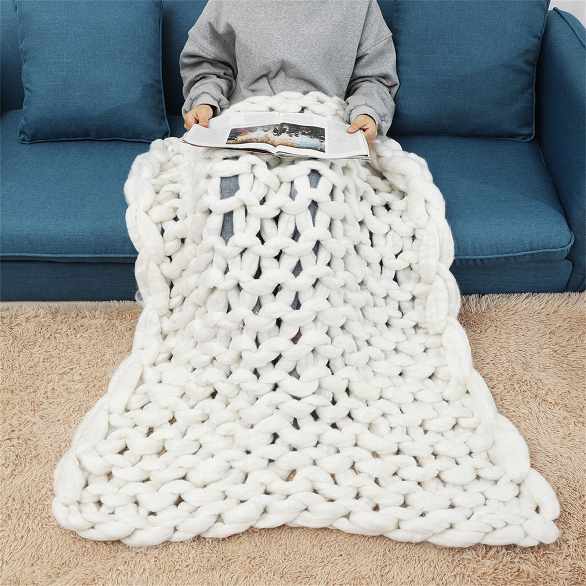 Person sitzt auf einem blauen Sofa, bedeckt mit einer weißen, grob gestrickten Decke.