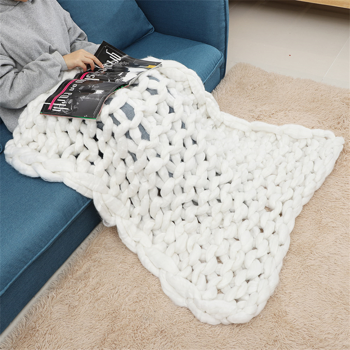 Eine Person sitzt auf einem blauen Sofa, bedeckt mit einer dicken, weißen Strickdecke, liest eine Zeitschrift.