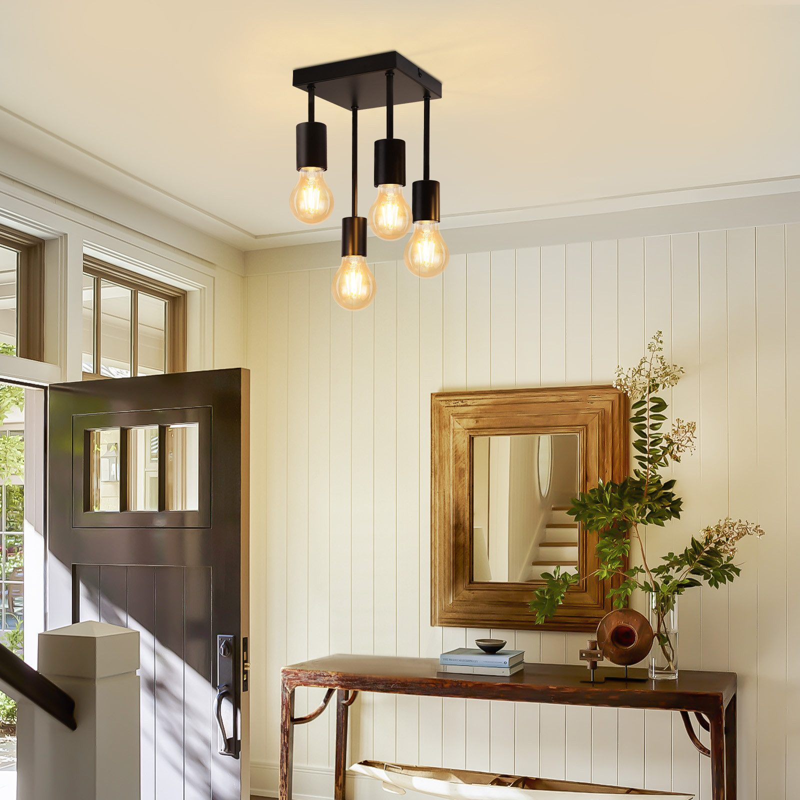 Wejście oświetlone czarną lampą sufitową z czterema wiszącymi żarówkami. Widoczne ciemne drzwi i stół z dekoracjami.