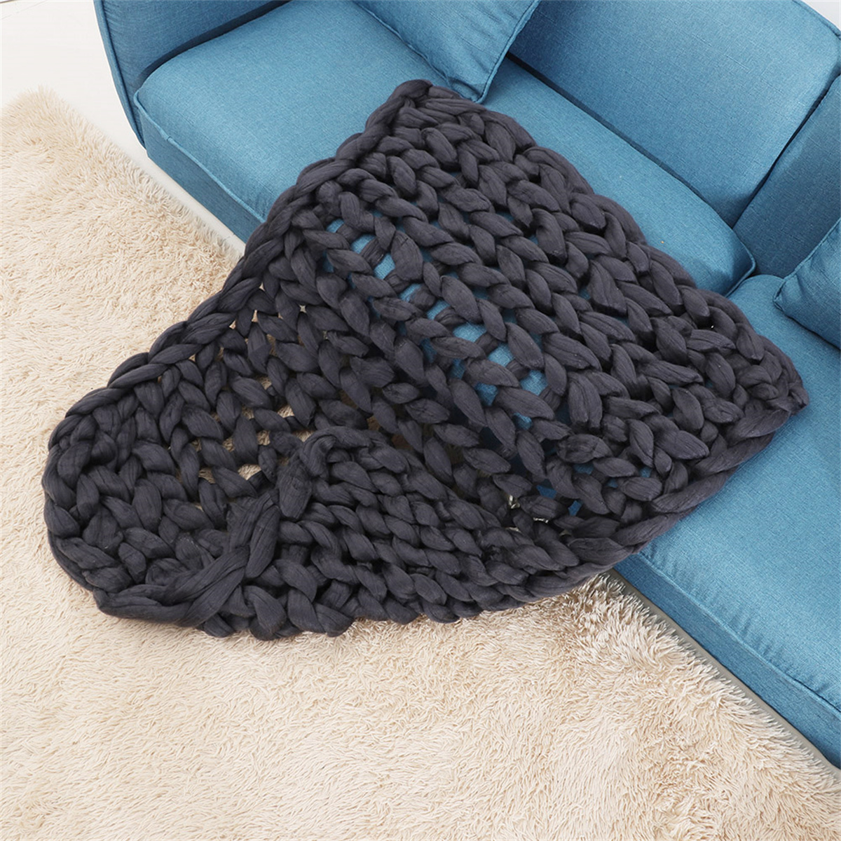 Eine dunkelgraue, grob gestrickte Decke liegt auf einem blauen Sofa auf einem beigen Teppich.