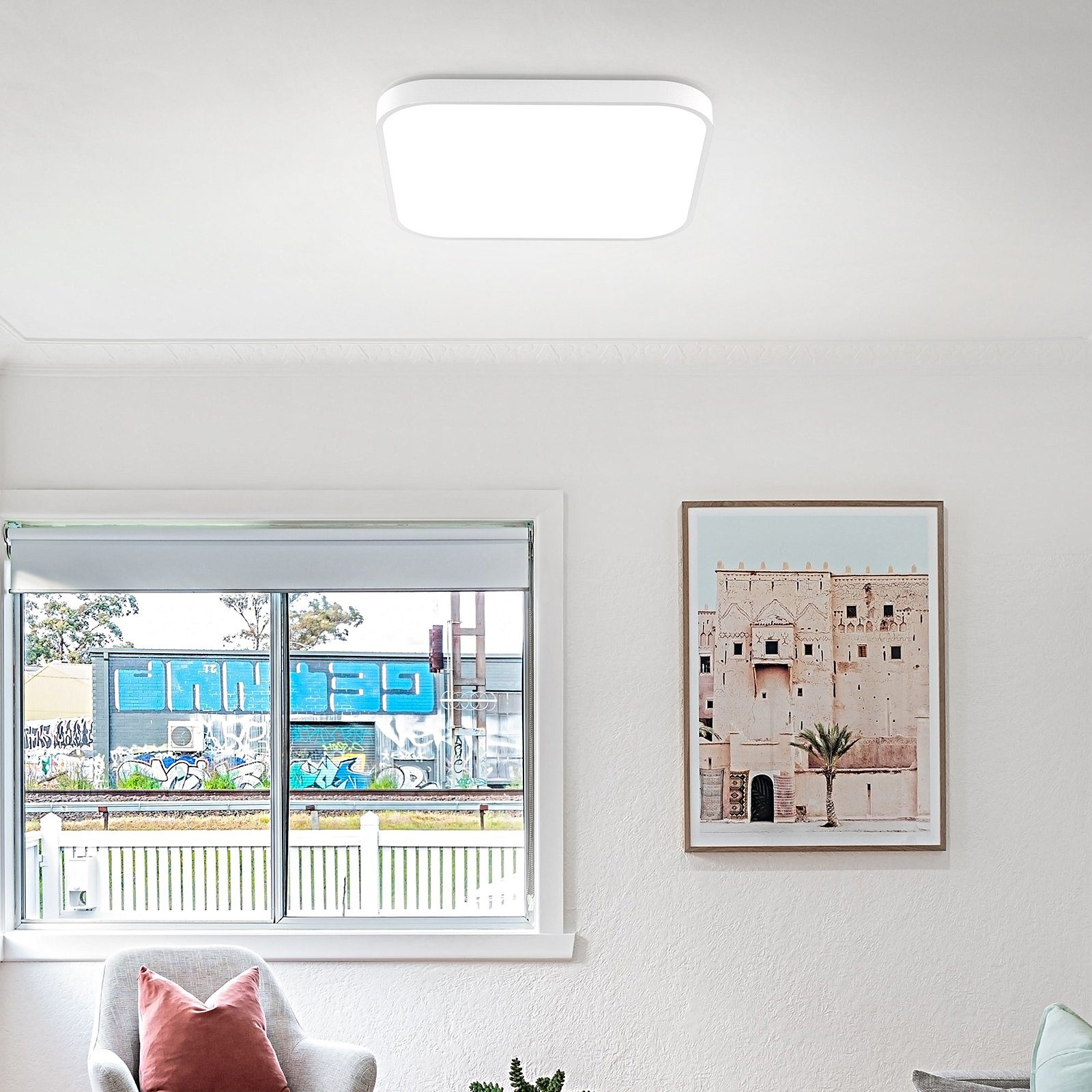 Pokój z lampą sufitową, oknem, obrazem i sofą. Ściana jest biała.