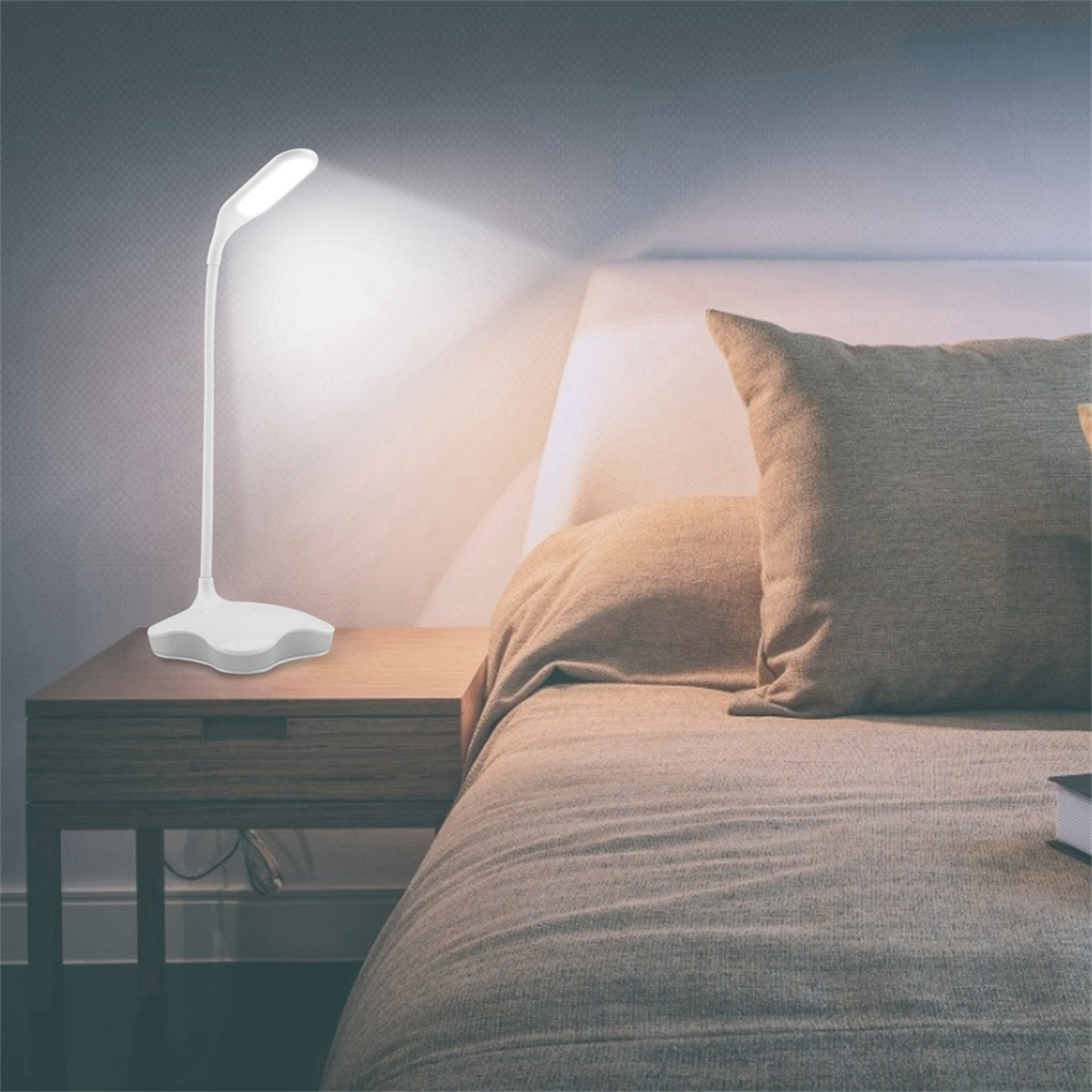 Eine weiße Schreibtischlampe auf einem Holztisch. Die Lampe ist an und beleuchtet ein Bett mit Kissen.