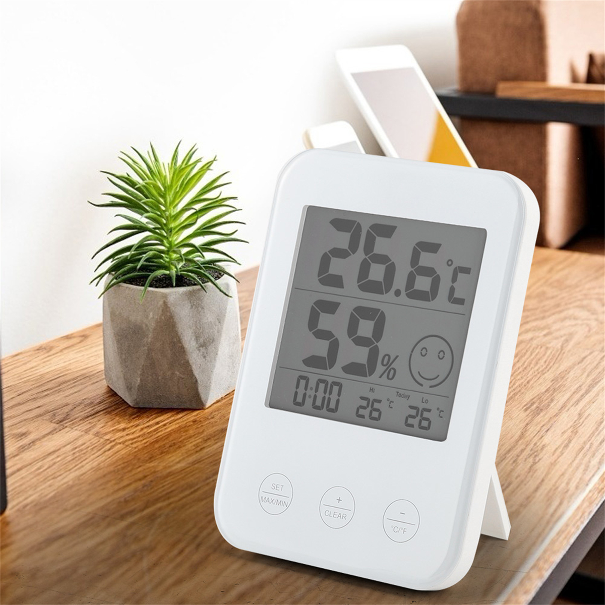 Weißes digitales Thermometer auf einem Holztisch, zeigt Temperatur und Luftfeuchtigkeit. Kleine grüne Pflanze.