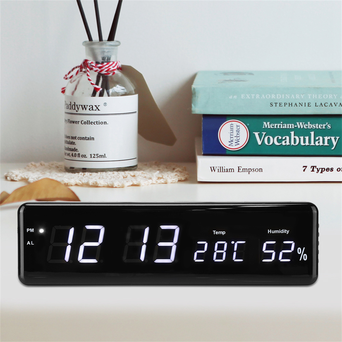 Schwarze Digitaluhr zeigt Zeit, Datum, Temperatur und Luftfeuchtigkeit. Bücher und eine Kerze sind im Hintergrund.