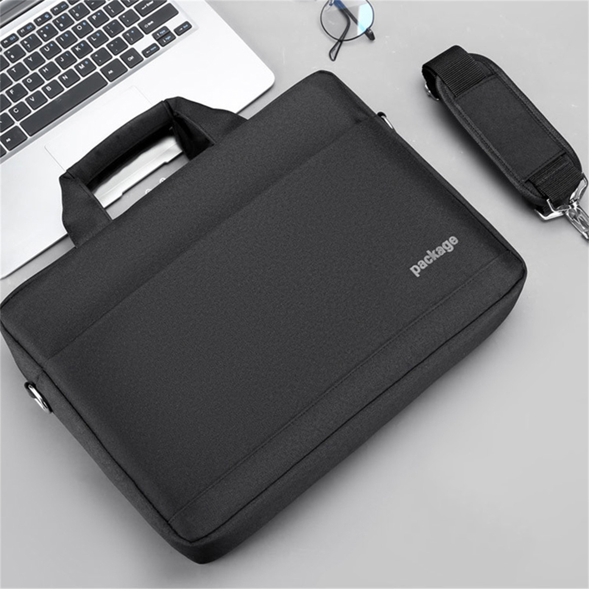 Eine schwarze Laptoptasche mit Griff und Schultergurt. Ein Laptop und eine Brille sind in der Nähe.
