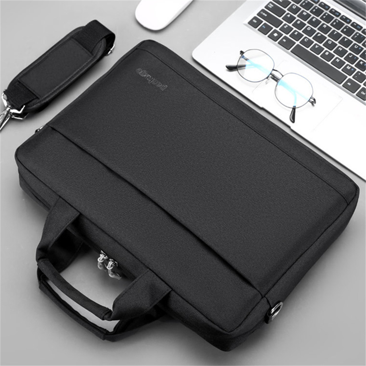 Schwarze Laptoptasche mit Griff, Schultergurt und Reißverschluss, auf grauer Oberfläche mit Laptop und Brille.