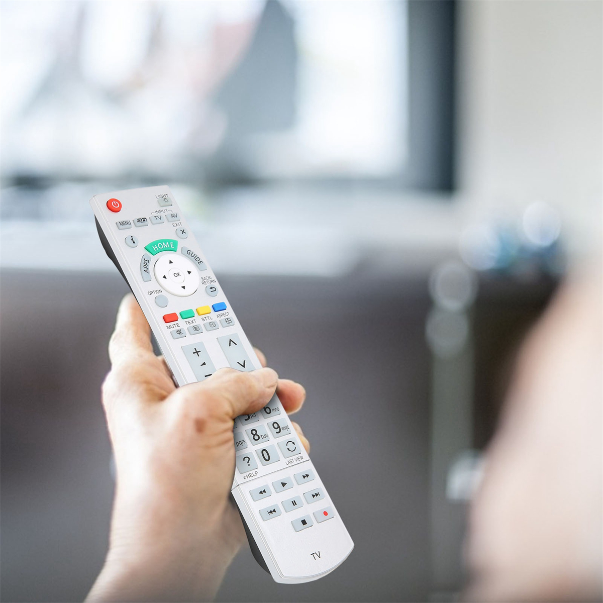 Hand, die eine weiße TV-Fernbedienung hält. Tasten umfassen Menü, Lautstärke und Kanalsteuerung. Fernseher im Hintergrund.