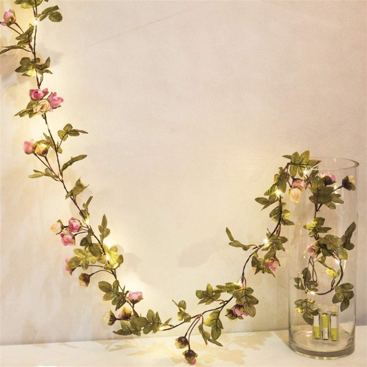 Eine Blumengirlande mit Lichtern und Blumen. Die Girlande ist an einer weißen Wand, neben einer Vase mit Lichtern.