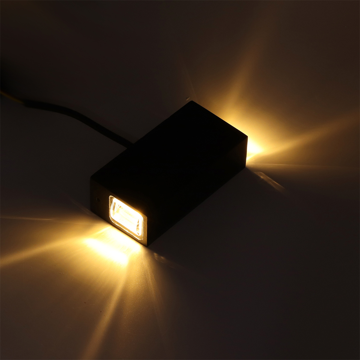 Schwarze rechteckige Wandleuchte, die gelbes Licht aussendet. Sie ist auf einer weißen Oberfläche montiert.