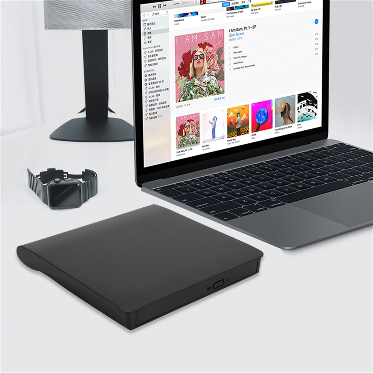 Laptop mit Musik-App, externe Festplatte und Smartwatch auf weißem Schreibtisch. Ein Lautsprecher im Hintergrund.