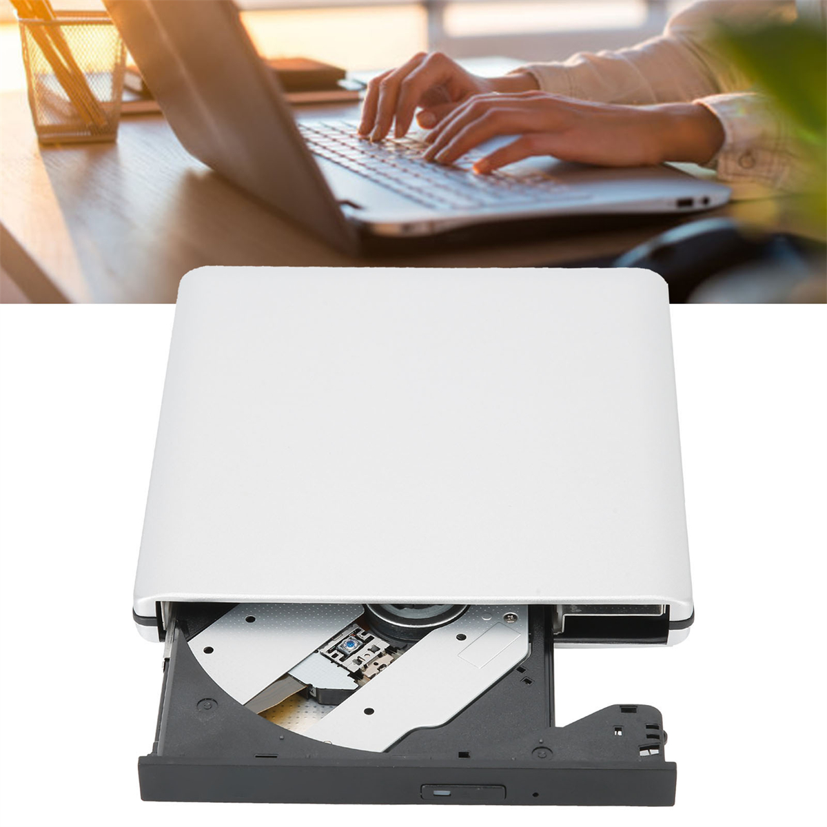 Hände tippen auf Laptop-Tastatur, ein weißes externes CD/DVD-Laufwerk im Vordergrund, auf einem Holzschreibtisch.