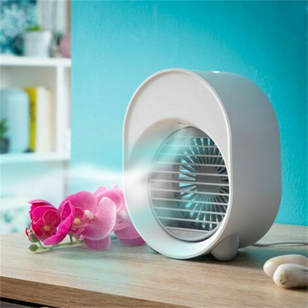 Ein weißer, ovaler Luftkühler mit Ventilator, rosa Blumen und ein Holztisch vor blauer Wand.