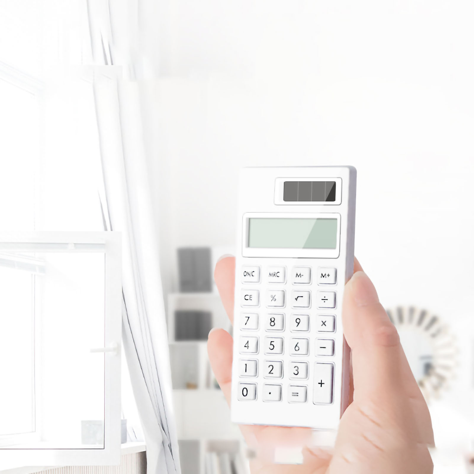 Ein weißer Taschenrechner in der Hand einer Person. Der Hintergrund ist ein verschwommenes Interieur mit Fenster.