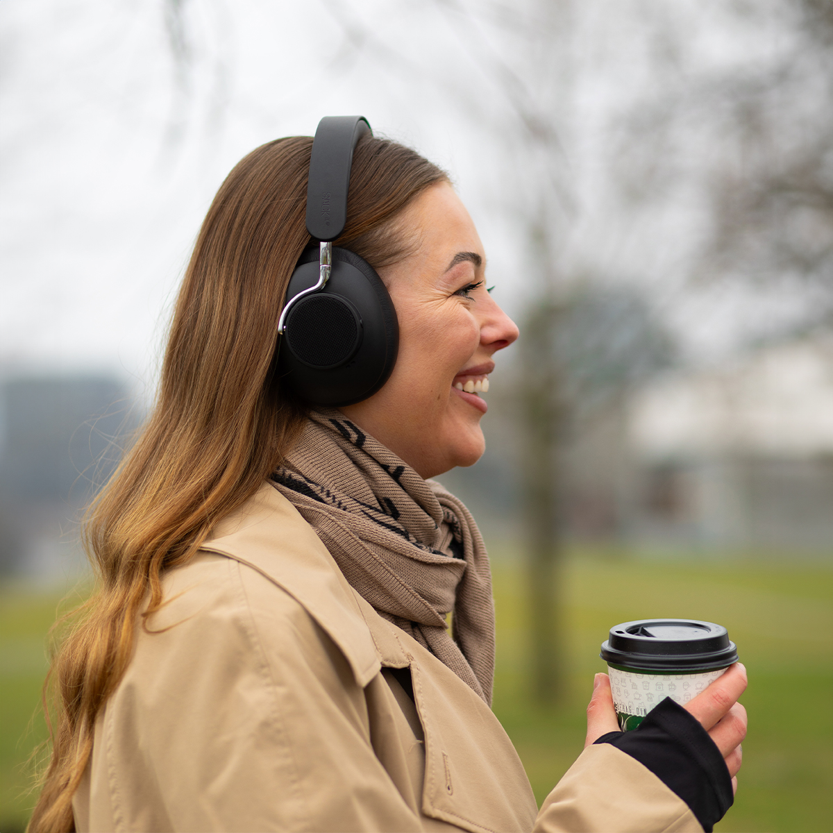 Una donna con cuffie e sciarpa che sorride, tiene una tazza di caffè, cammina all'aperto.