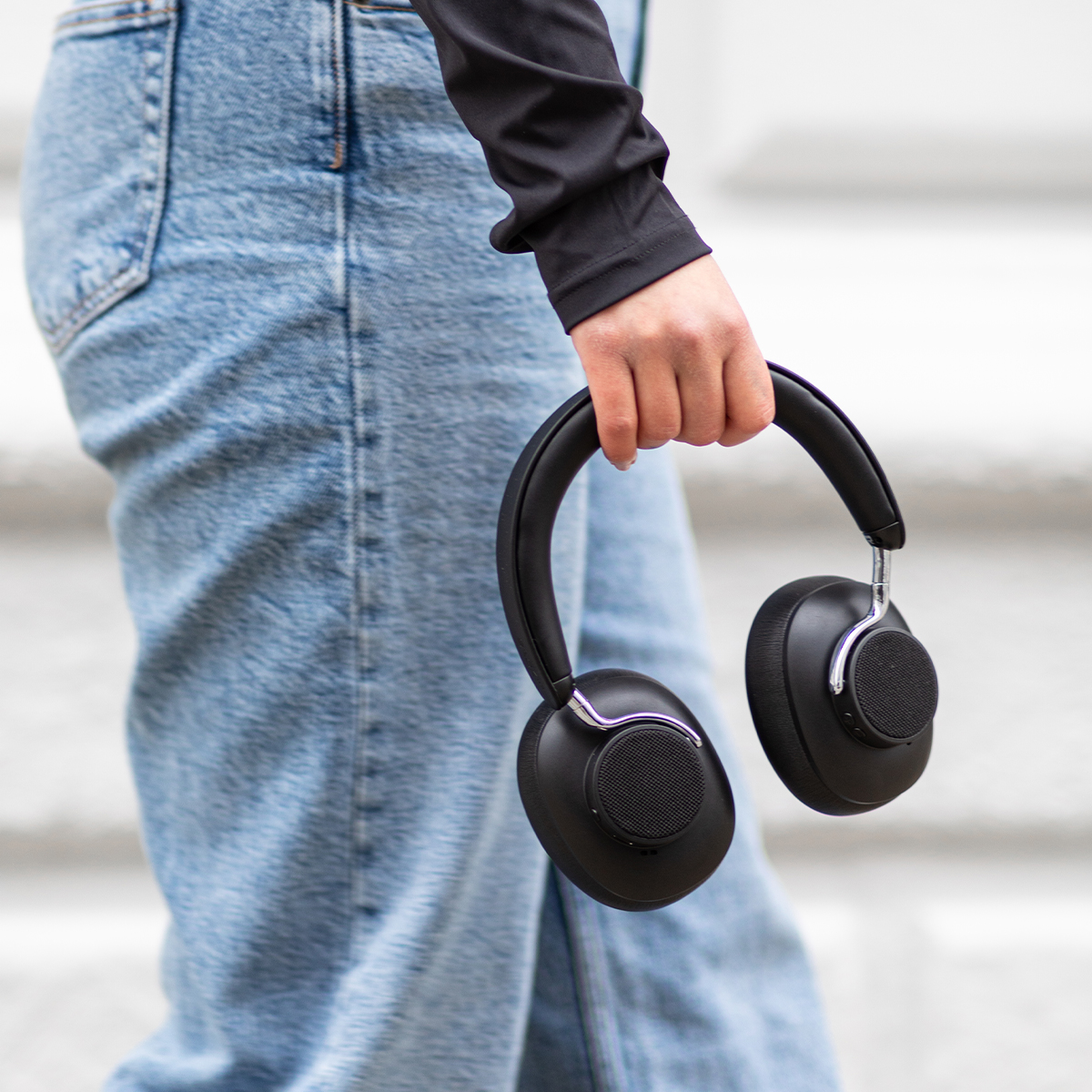 Persona che tiene cuffie nere, indossa jeans blu e una camicia nera. Sfondo bianco.