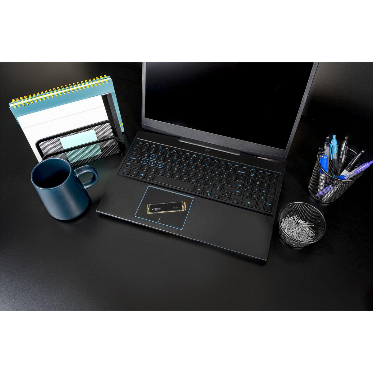 Laptop, kalendarz, kubek i materiały biurowe na czarnym biurku. Dysk SSD jest na laptopie.