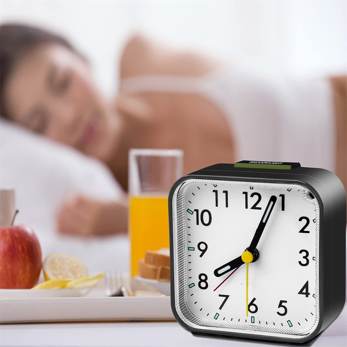 Wecker auf Tisch. Person im Bett unscharf im Hintergrund. Frühstückstablett mit Essen.