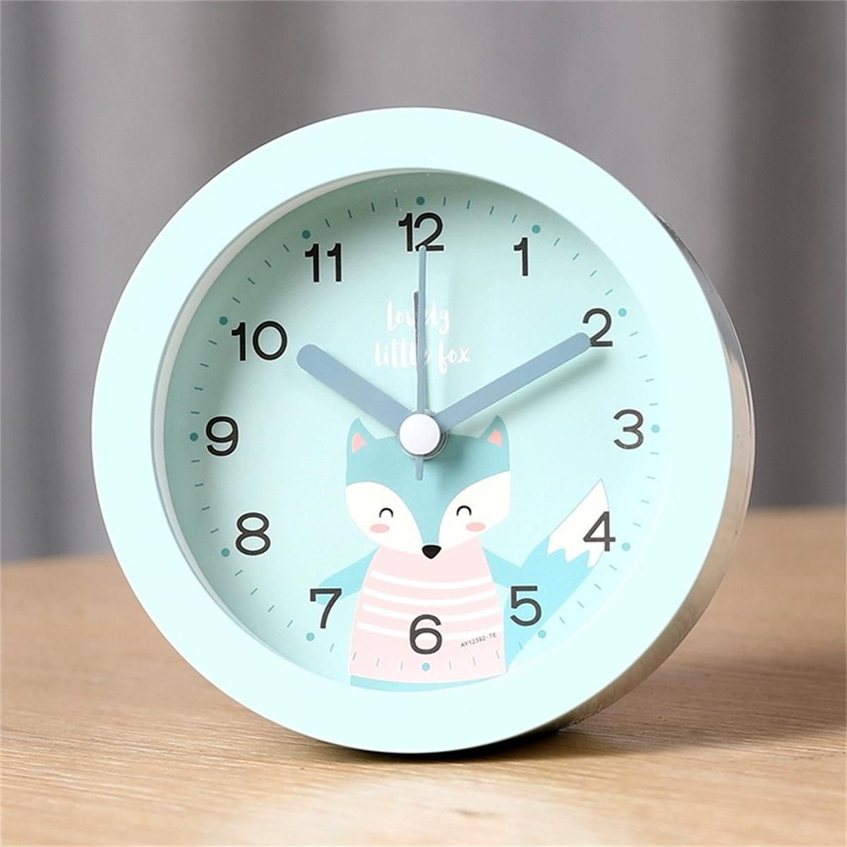 Ein hellblauer Wecker mit Fuchsmotiv. Er hat ein rundes Zifferblatt mit Zahlen und Zeigern. Die Uhr steht auf einem Tisch.
