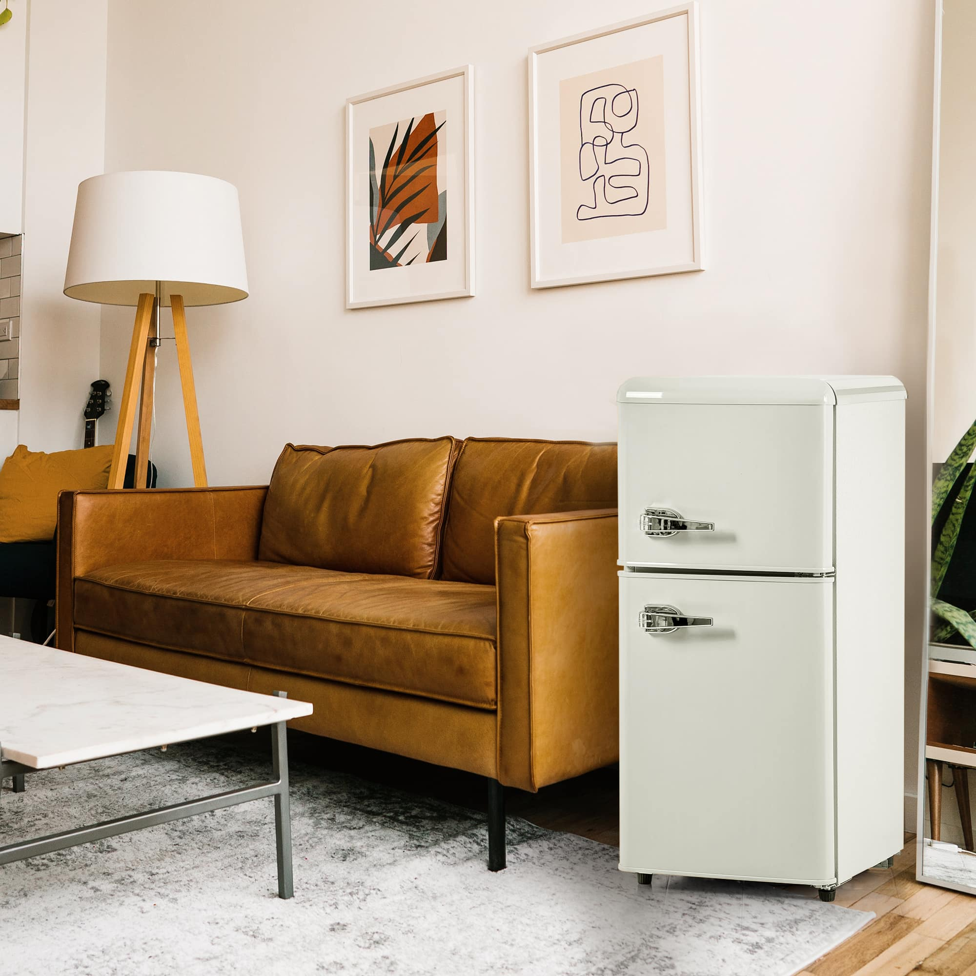 Ein kleiner weißer Kühlschrank steht in einem Wohnzimmer. Eine Ledercouch und Kunstwerke sind im Hintergrund.