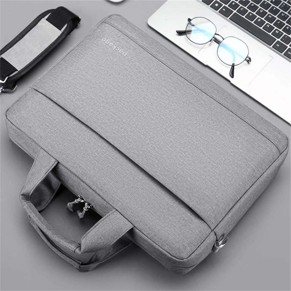 Graue Laptoptasche mit Griff, Riemen und einem Laptop, Brille auf grauer Oberfläche.