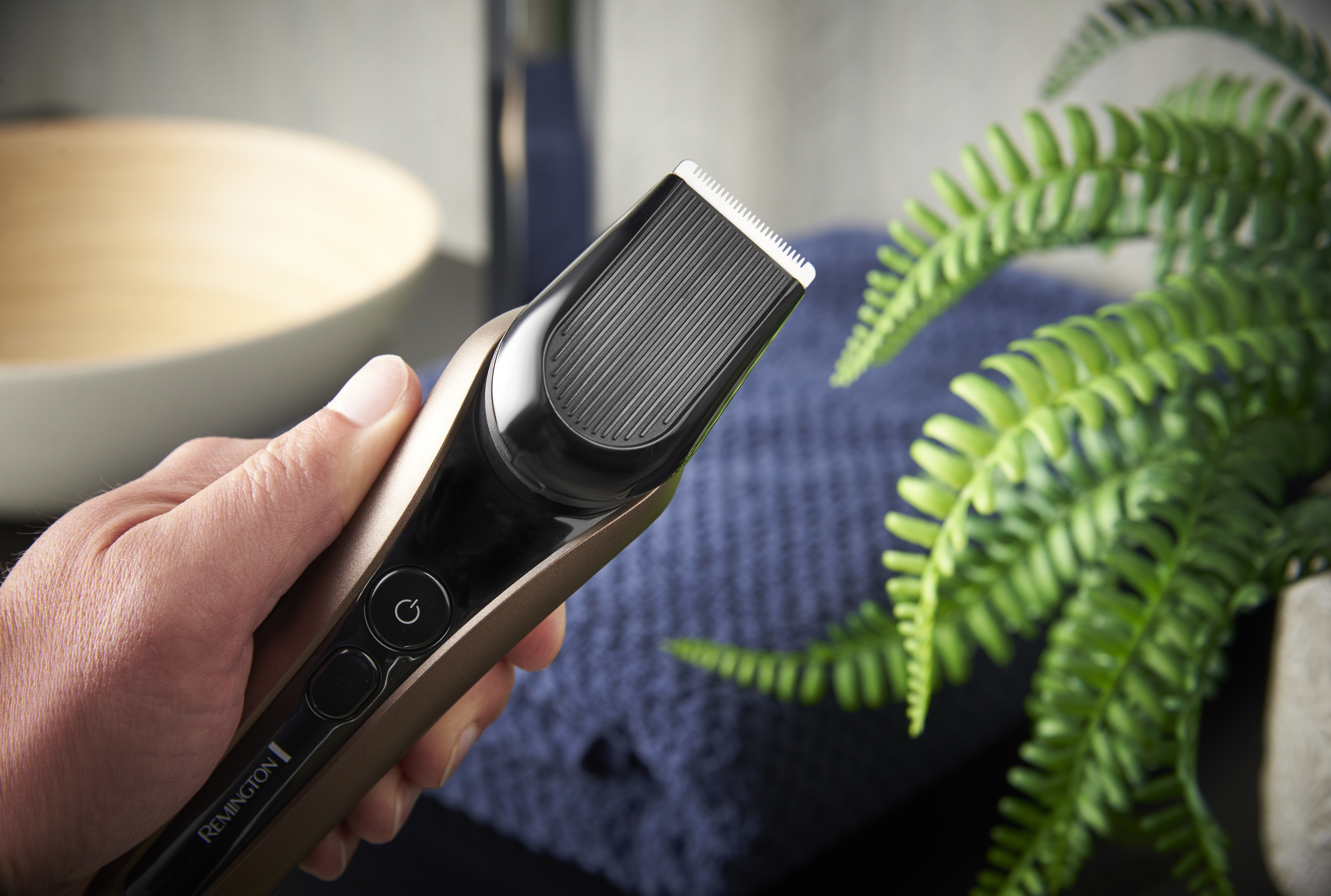 Eine Hand hält einen gold-schwarzen Trimmer. Farn und ein blaues Handtuch im Hintergrund.