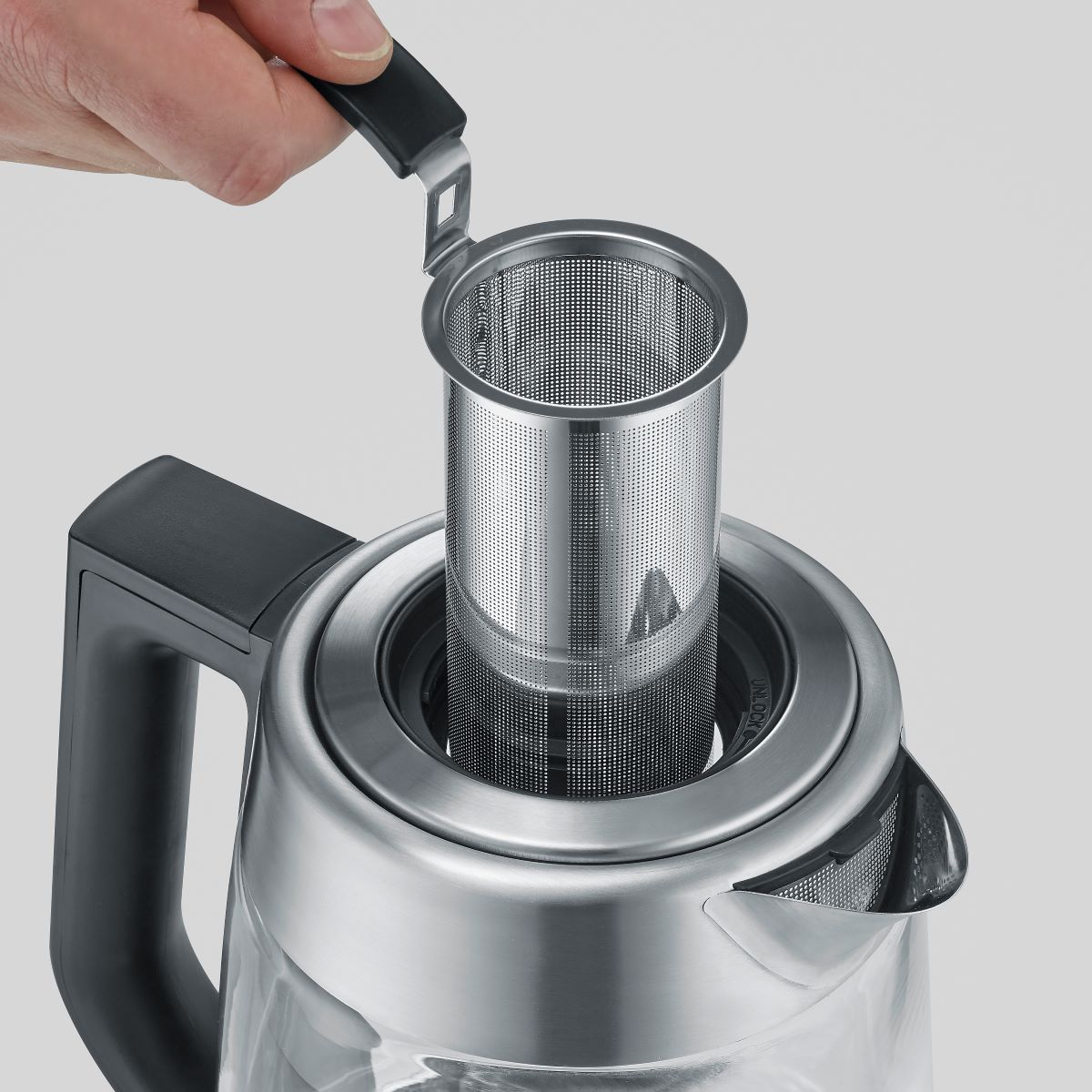 Een hand die een thee-infuser in een glazen waterkoker plaatst. De waterkoker is helder met een zwart handvat.