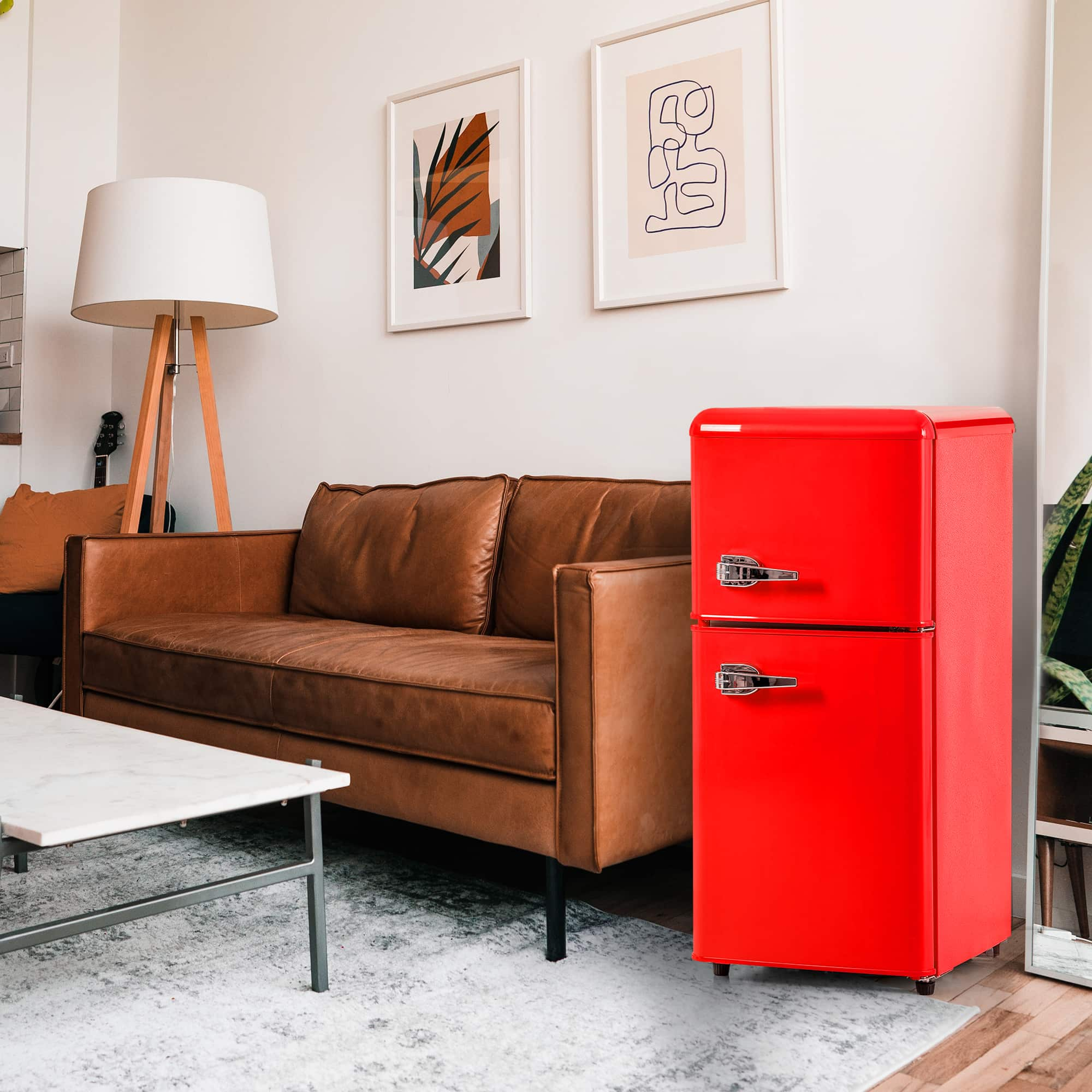 Ein roter Retro-Mini-Kühlschrank steht neben einem braunen Ledersofa, mit Couchtisch und Kunst im Hintergrund.