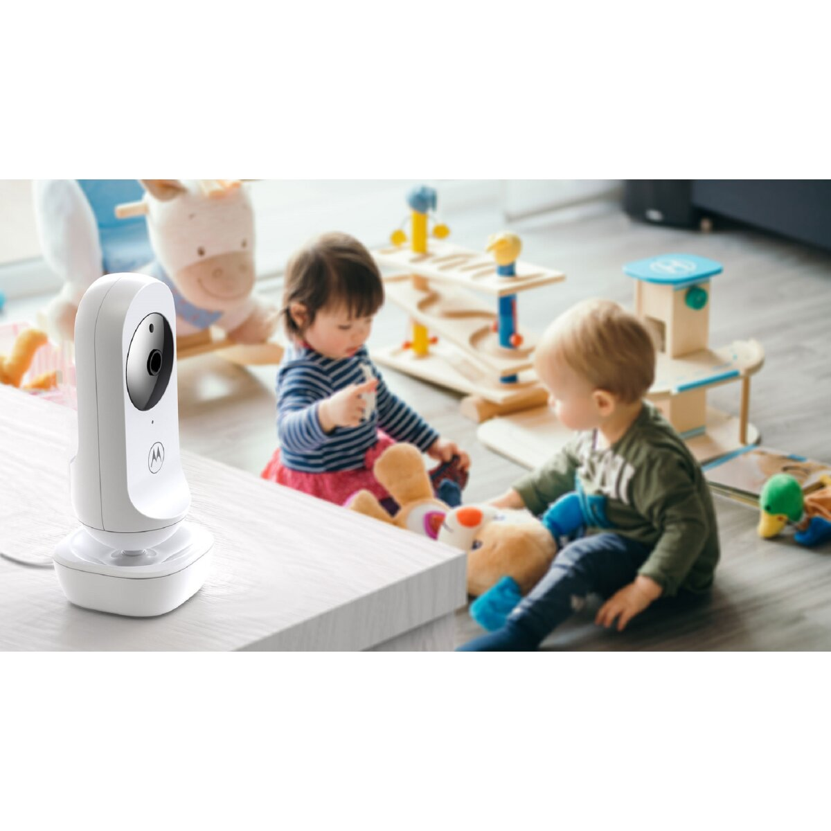 Ein weißer Babyphone auf einem Tisch. Zwei Babys spielen im Hintergrund mit Spielzeug.