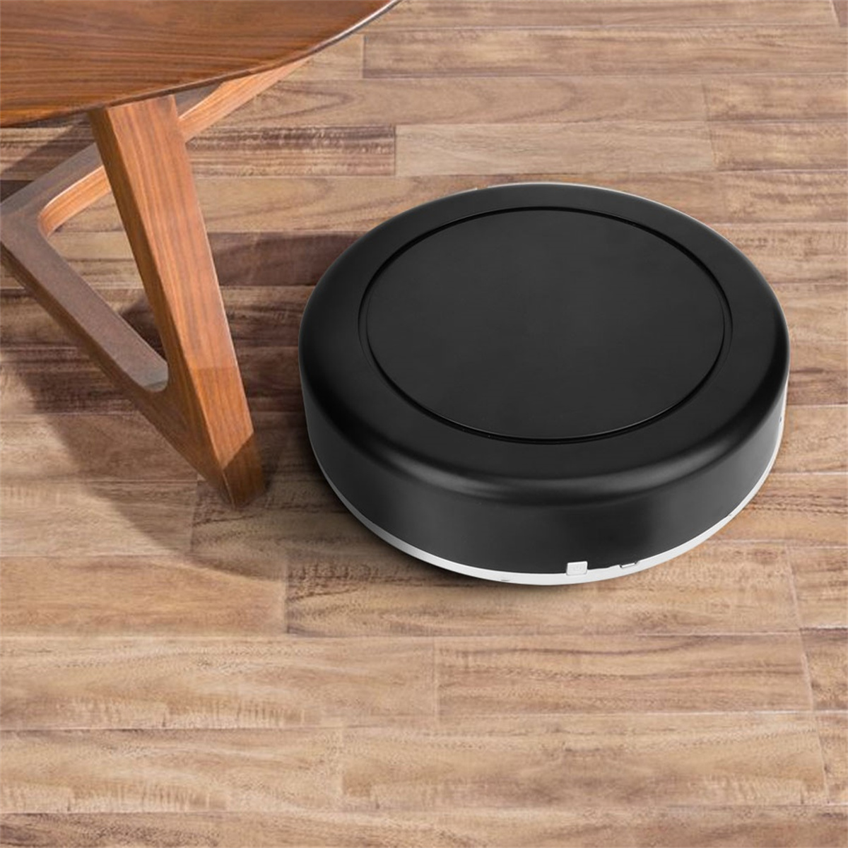 Schwarzer runder Roboter-Staubsauger auf Holzboden in der Nähe eines Tischbeins.