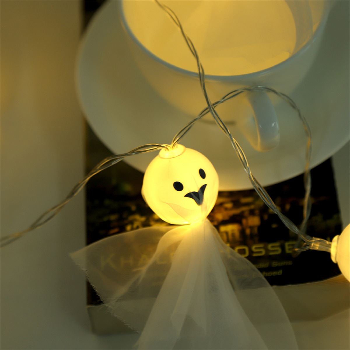 Eine Lichterkette mit einer geisterförmigen Glühbirne. Die Glühbirne leuchtet, auf einem Buch, mit einer Tasse im Hintergrund.