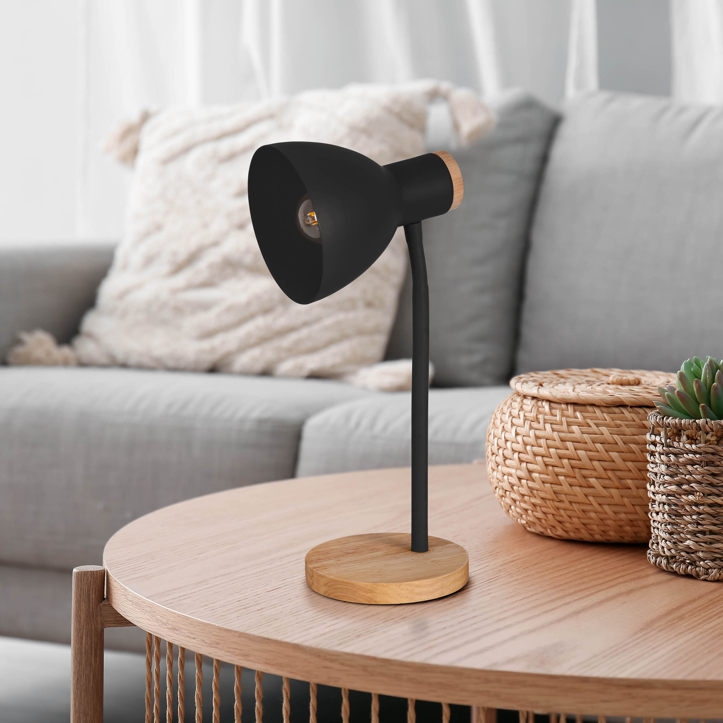 Eine schwarze Schreibtischlampe auf Holzsockel. Sie steht auf einem Holztisch neben einem grauen Sofa und einer Pflanze.