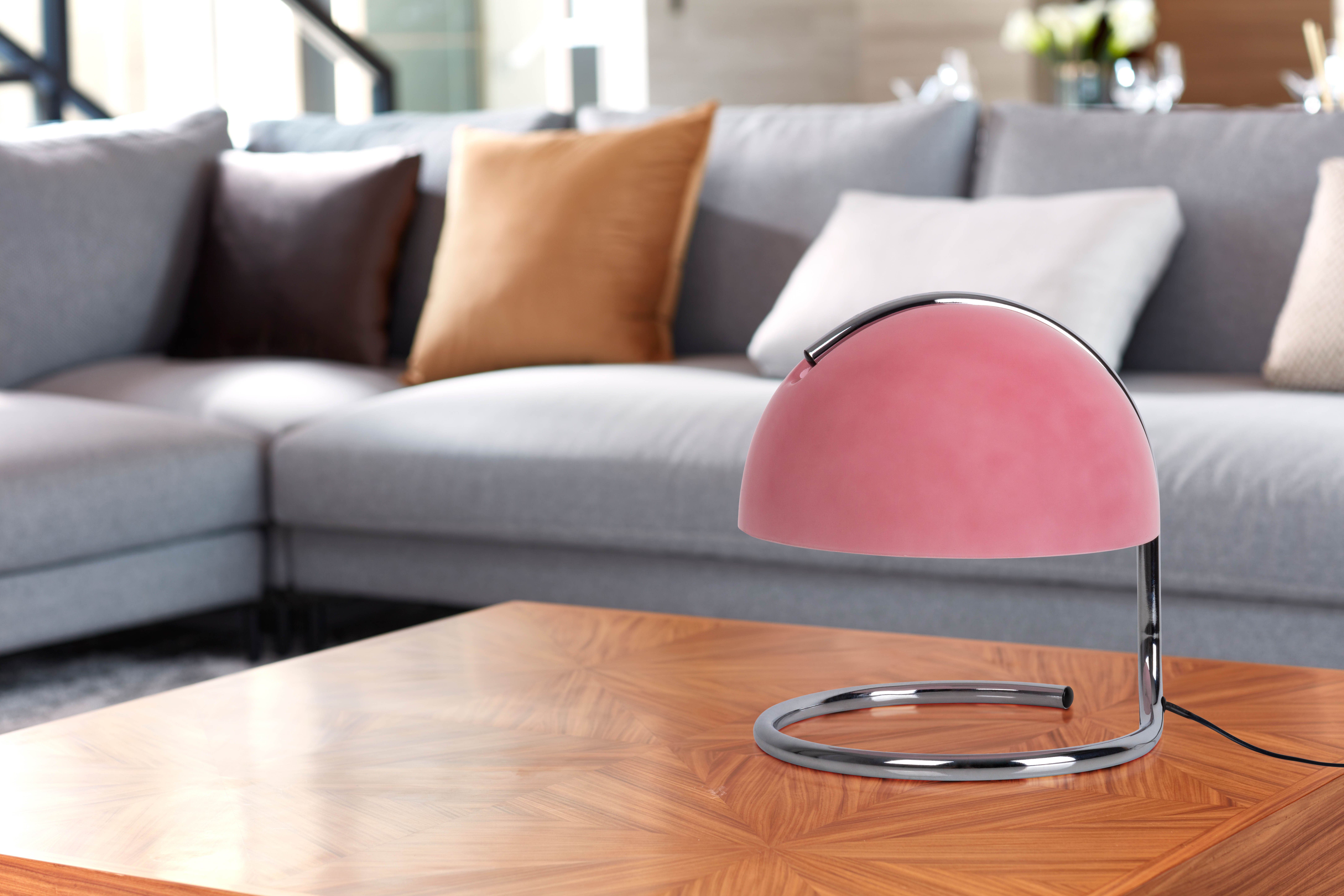 Rosa Lampe auf einem Holztisch. Der Tisch steht vor einer grauen Couch und Kissen.