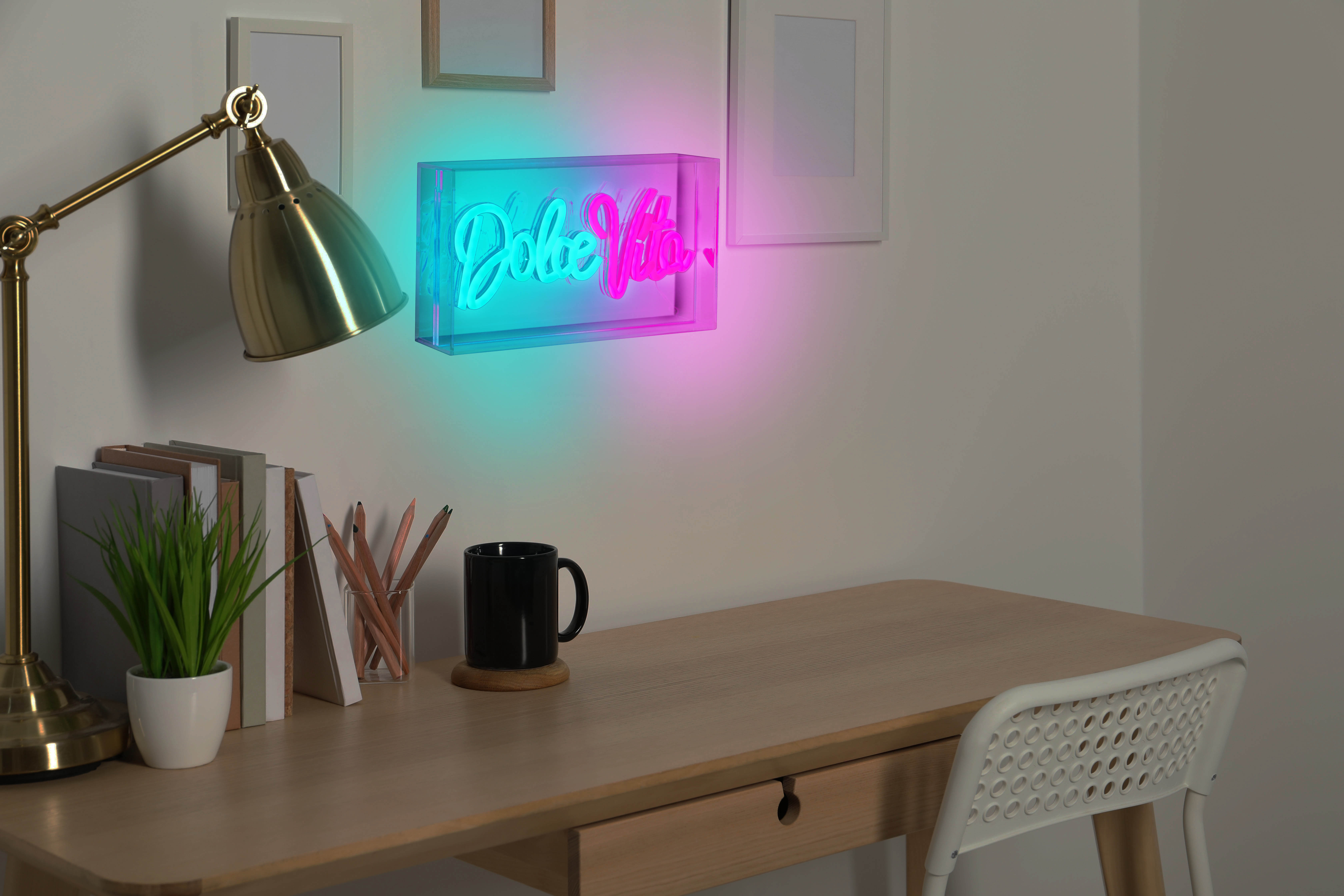 Schreibtischszene mit Neon-Schild, Lampe, Büchern und einer Tasse. Wandkunst und ein Stuhl sind ebenfalls sichtbar.