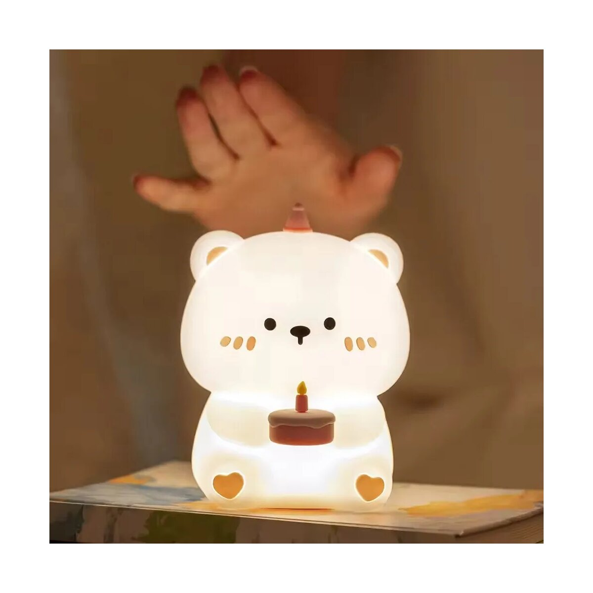 Ein weißes bärenförmiges Nachtlicht mit Kuchen und einer Kerze. Eine Hand ist darüber. Der Hintergrund ist unscharf.