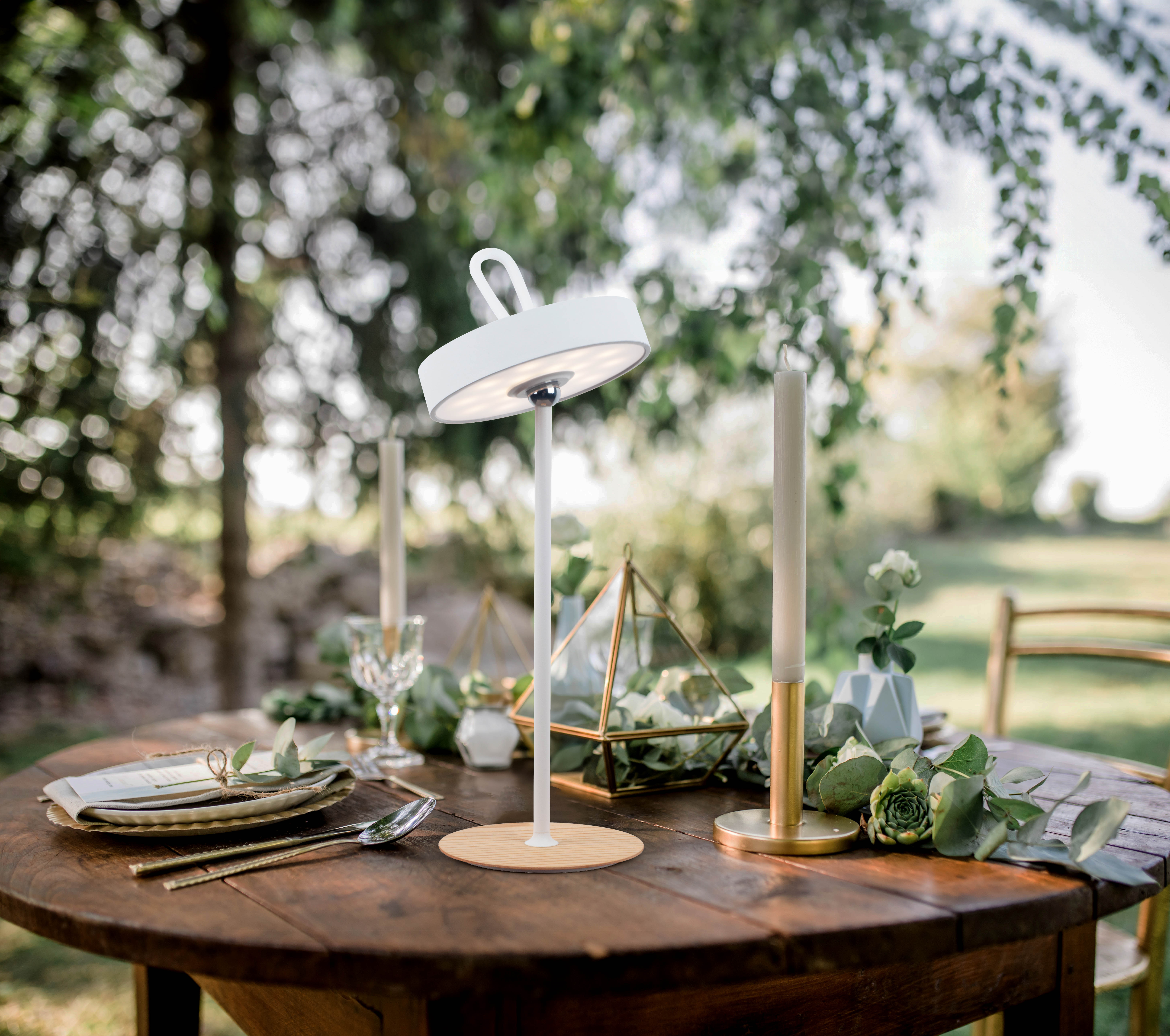 Eine weiße Lampe auf einem Holztisch. Kerzen, Blumen und ein Glas stehen ebenfalls auf dem Tisch. Bäume sind im Hintergrund.