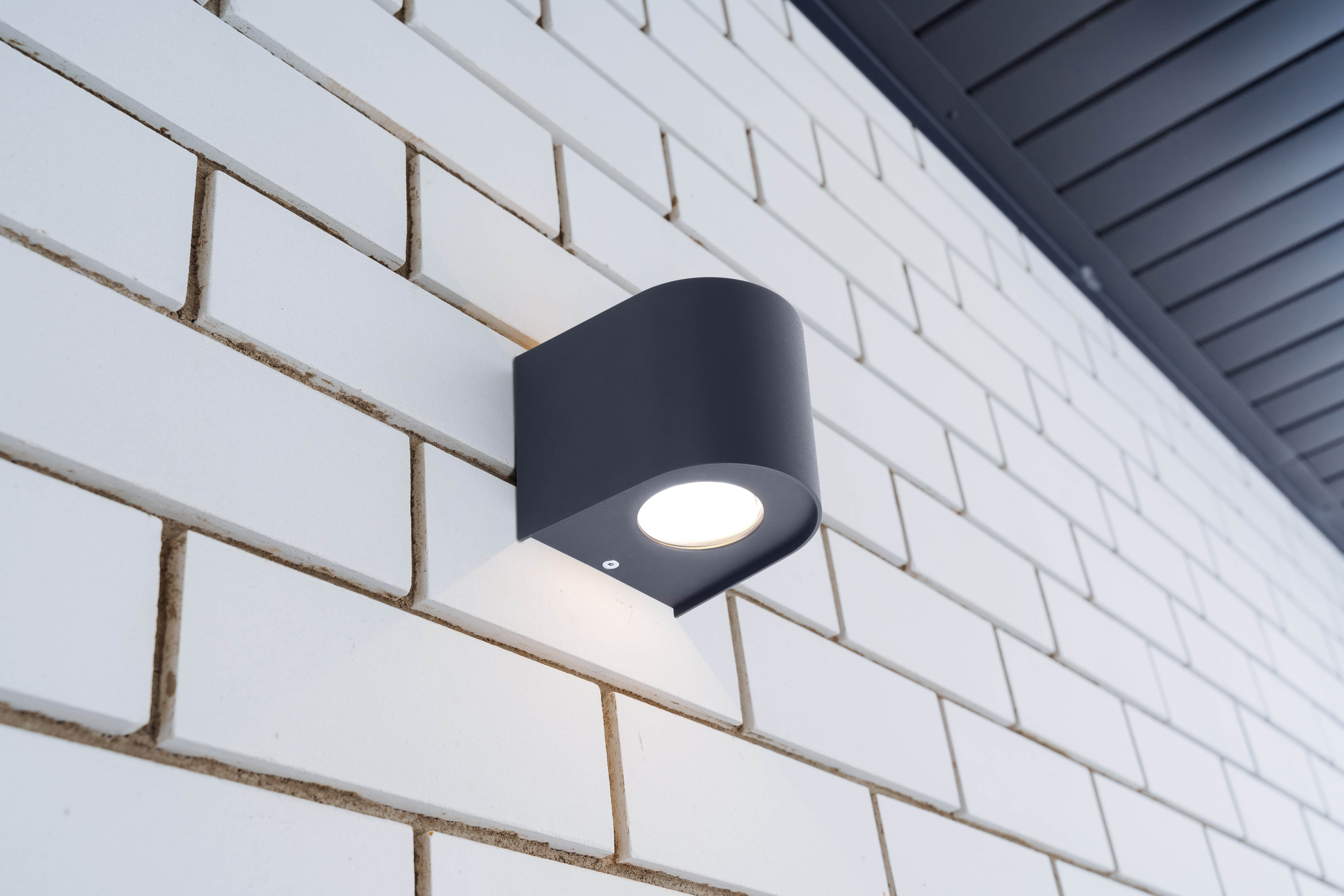 Schwarze Außenwandleuchte an einer weißen Backsteinmauer. Die Lampe hat ein modernes, abgerundetes Design und gibt Licht ab.