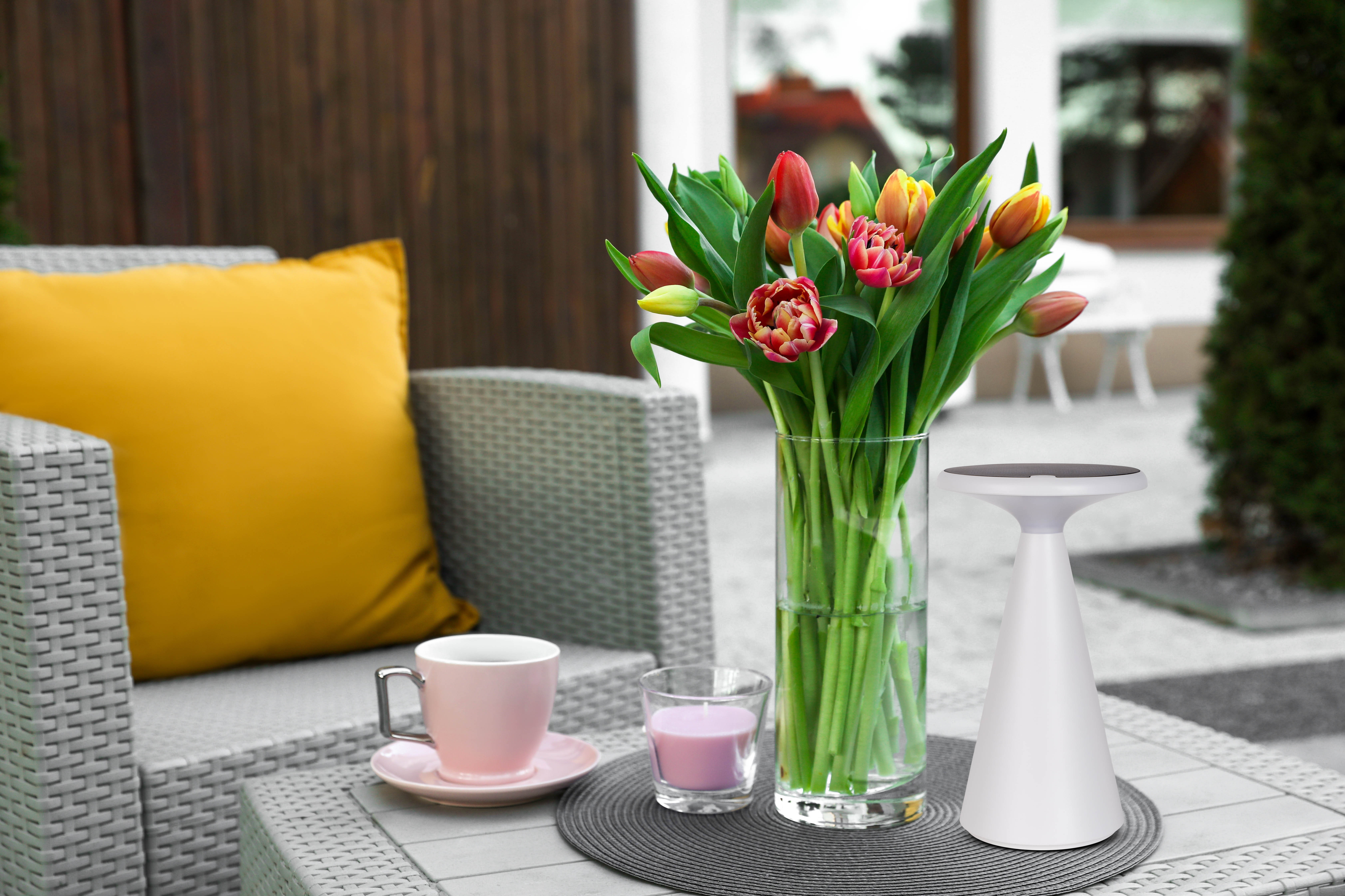 Eine Terrasse mit Blumen in einer Vase, einem Tisch mit Tasse und Kerze und einem gelben Kissen.