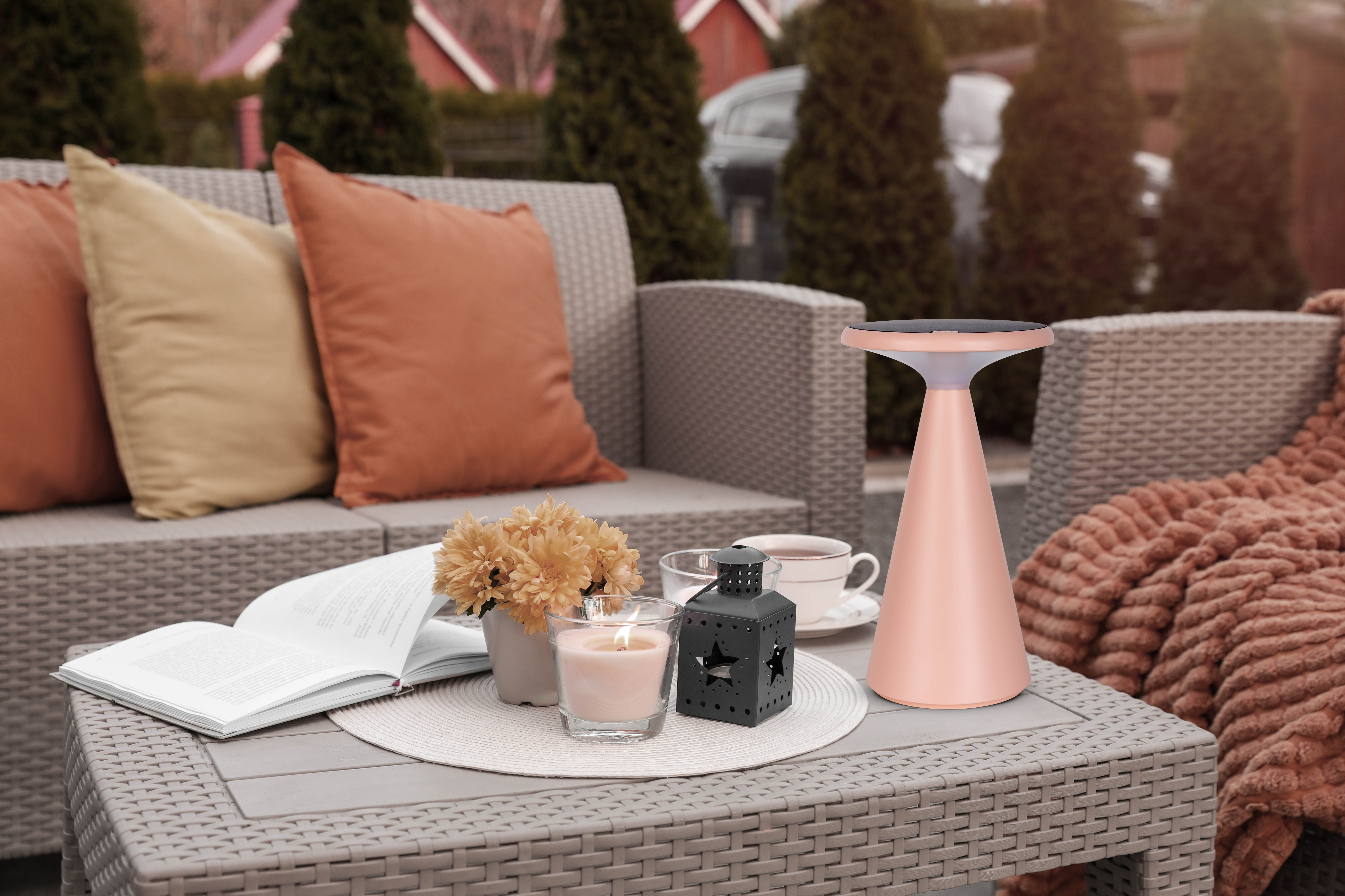Szene im Freien mit Tisch und Möbeln. Auf dem Tisch befinden sich eine Lampe, eine Kerze, ein Buch, Blumen und eine Decke.